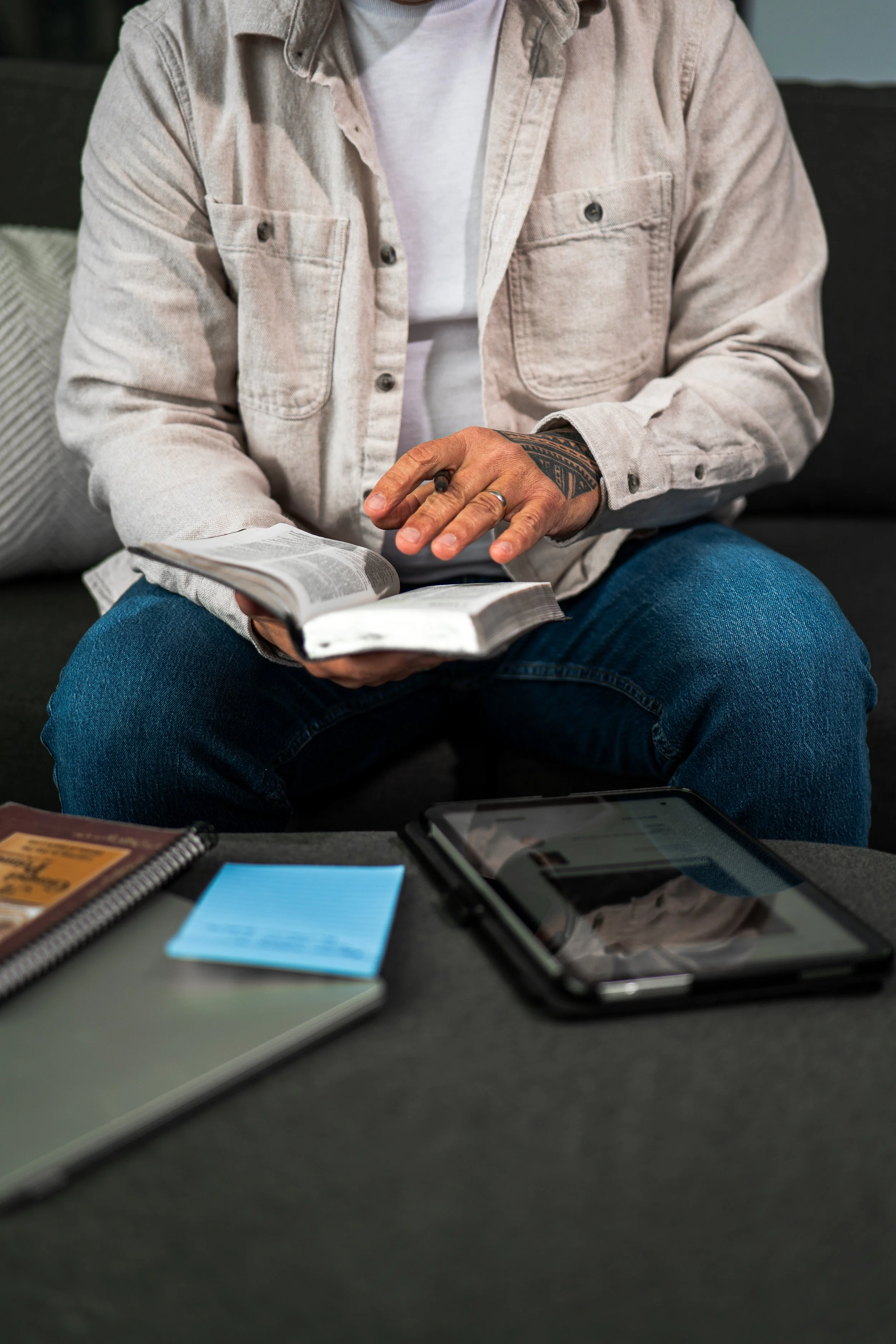 Person wearing a beige jacket with tattoos on their left hand, holding an open Bible or book, sitting on a couch with a smartphone and notebook on the table in front of them.