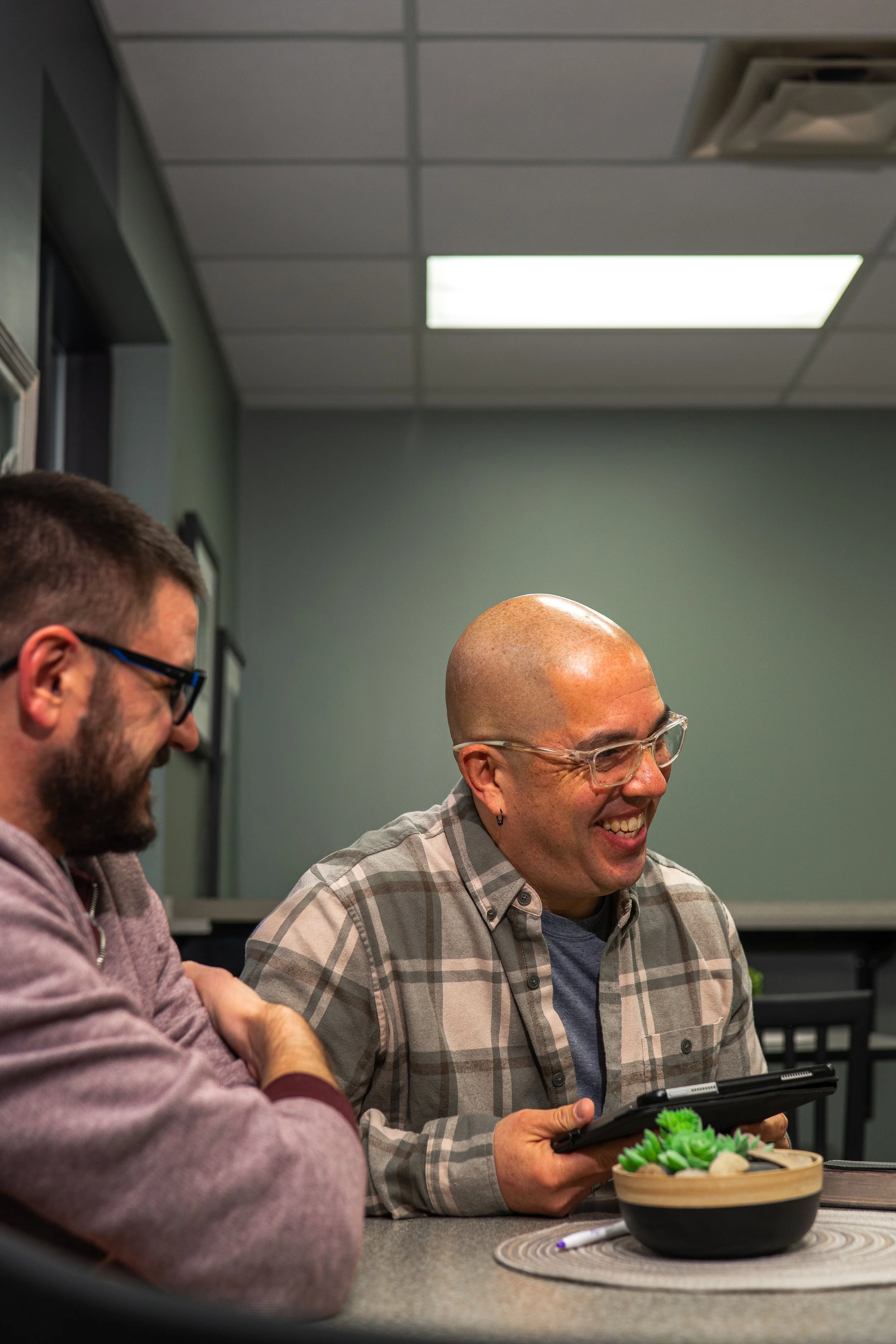 Two men sitting at a table, looking at a tablet, laughing. One man is bald with glasses and earrings, the other has dark hair and a beard, wearing glasses. There is a bowl of green succulents on the table.