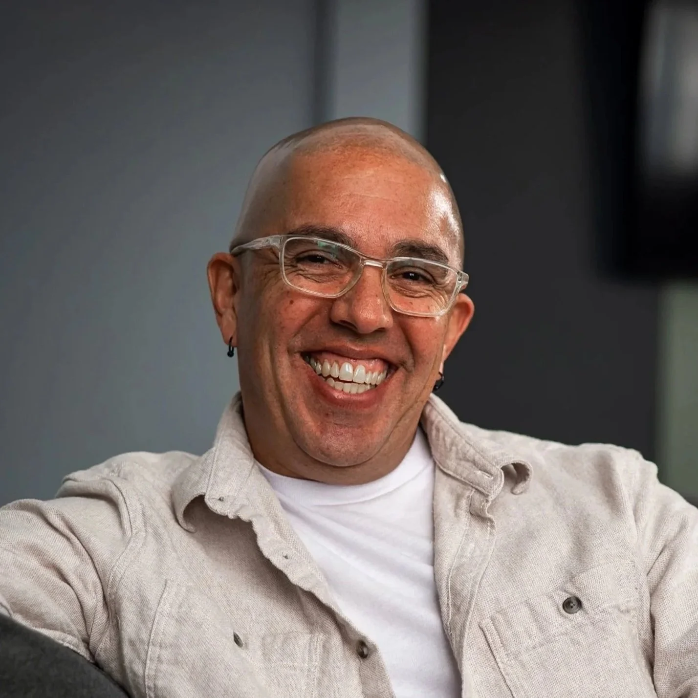A smiling man with glasses, earrings, and a bald head standing outdoors in front of a brick wall.