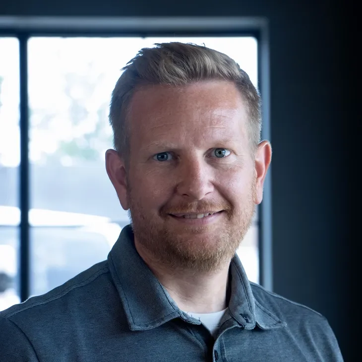 A man with light skin, short blond hair, and a beard, smiling, wearing a gray collared shirt, indoors with large windows in the background.