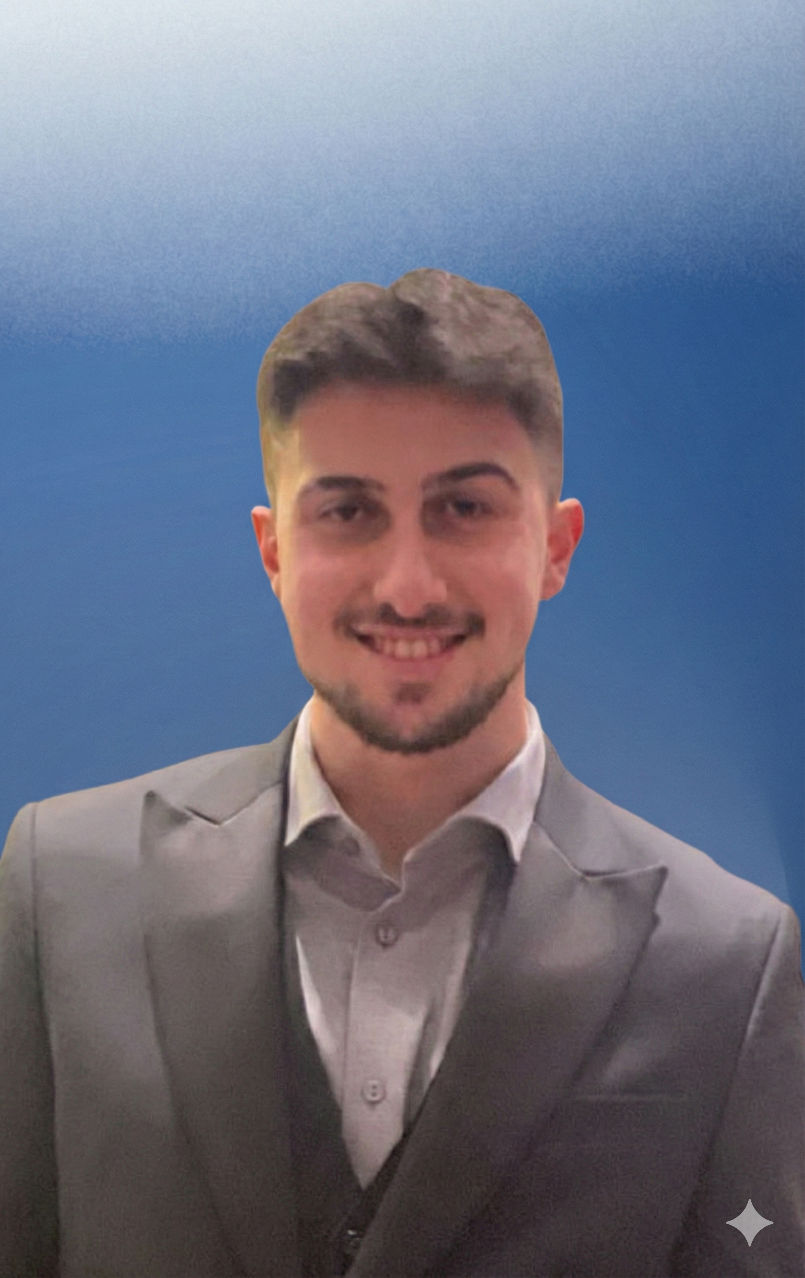 A young man with short brown hair, a trimmed beard, dressed in a light gray suit and white shirt, smiling against a gradient blue background.