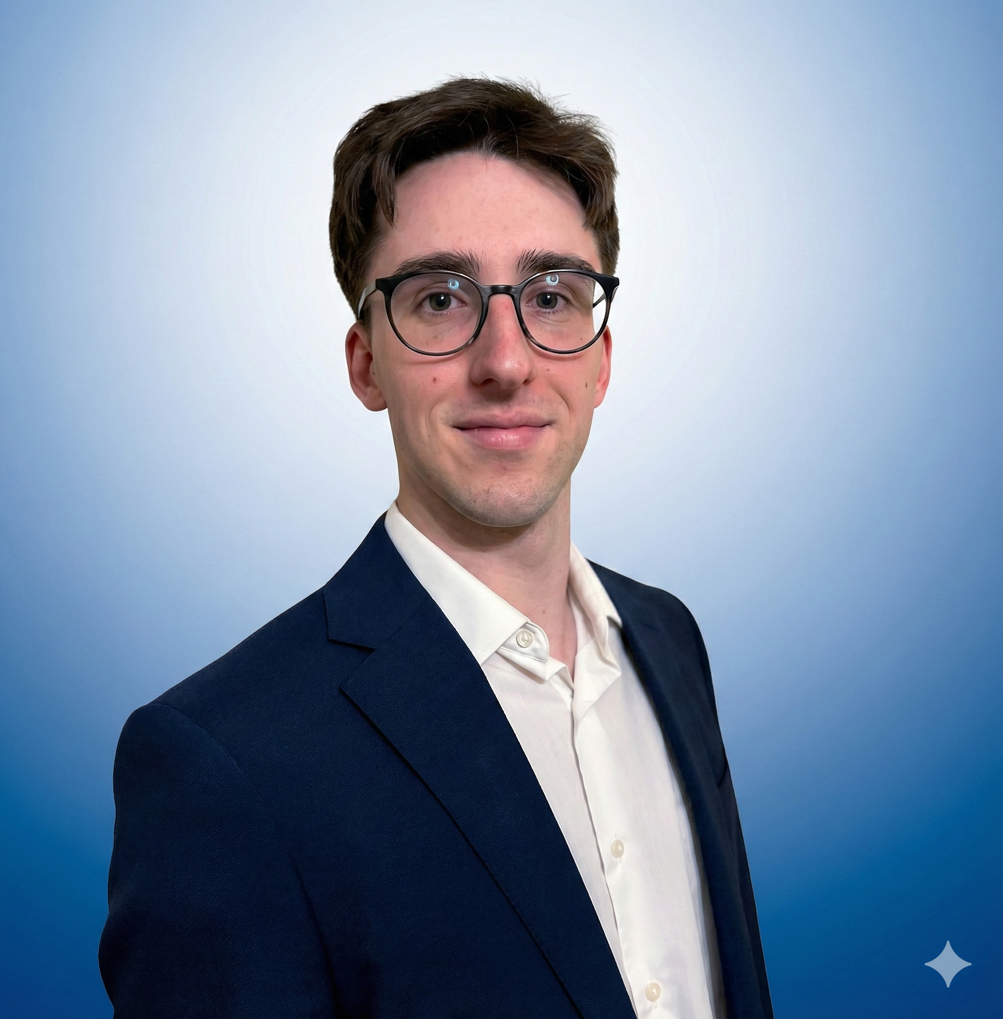 A young man wearing glasses in a navy blazer and light dress shirt, smiling at the camera against a gradient blue background.
