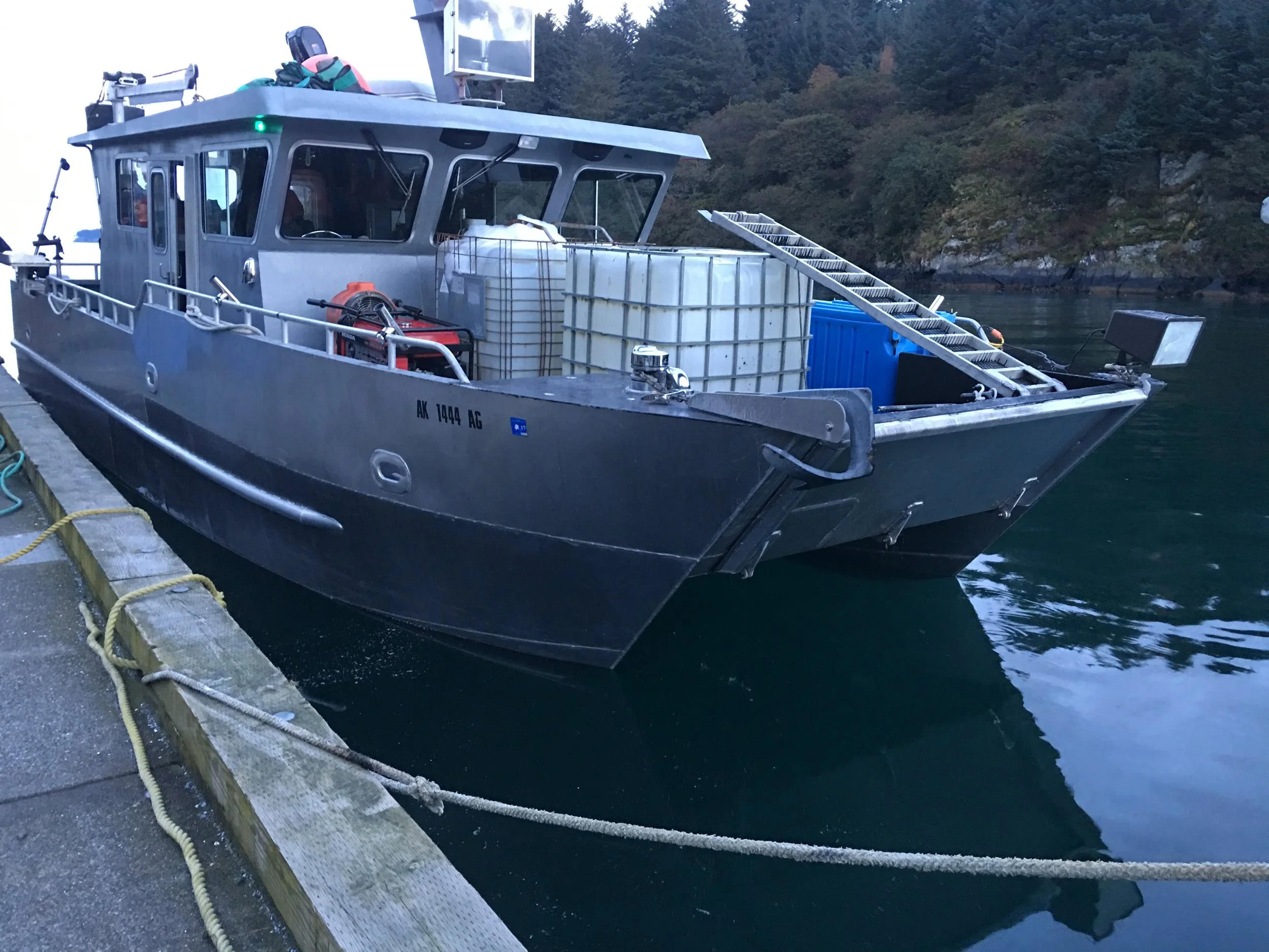 A gray work boat docked at a pier with storage tanks, equipment, and an extendable ladder, surrounded by water and forested hills in the background.