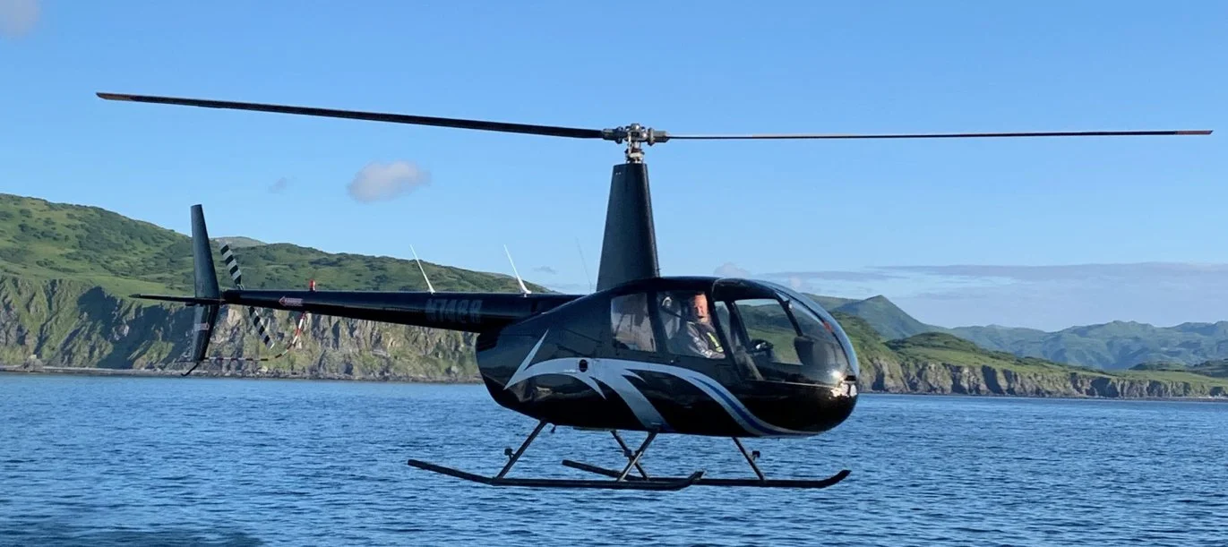 A black helicopter flying over a body of water with green hills and mountains in the background.