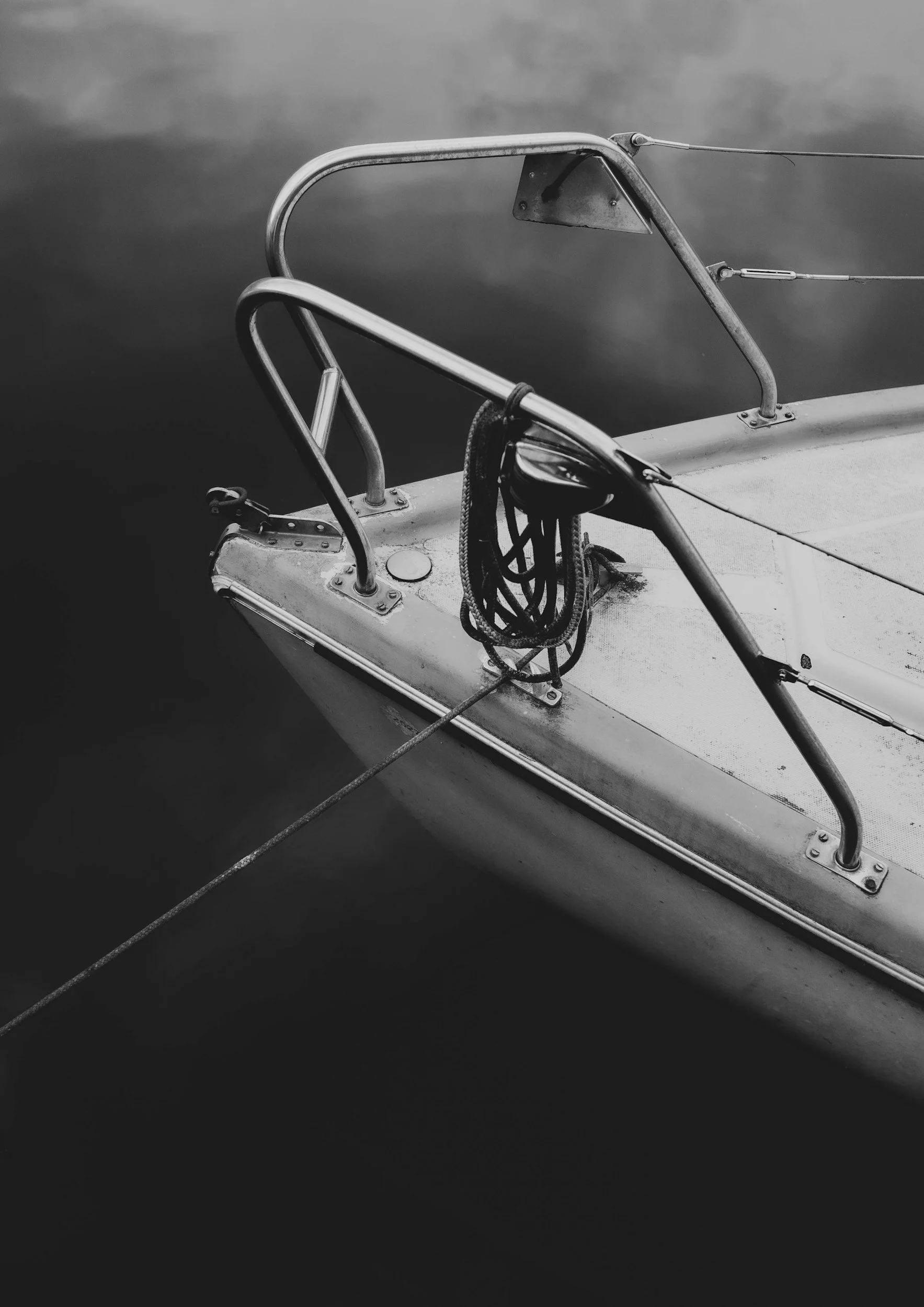 Close-up of a sailboat's bow with metal railings and a coiled rope, floating on calm water in black and white.