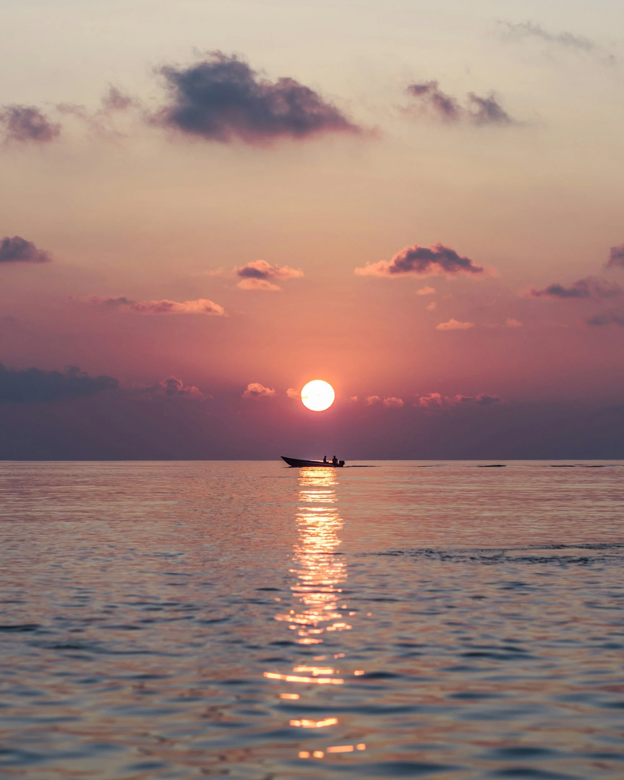 Sunset over calm ocean with a silhouetted boat and a few people on board, pink and purple clouds in the sky.