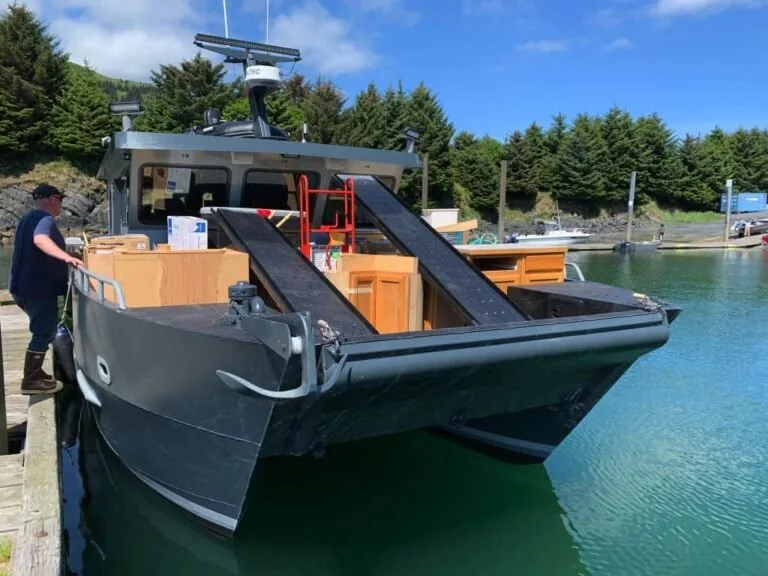 A large black boat docked at a marina with a man on the dock. The boat has two slides on the deck and various boxes and equipment. The scene is set on a sunny day with trees and a blue sky in the background.