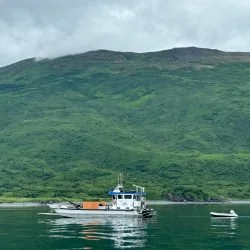 A boat on calm water with green hills in the background under cloudy sky.