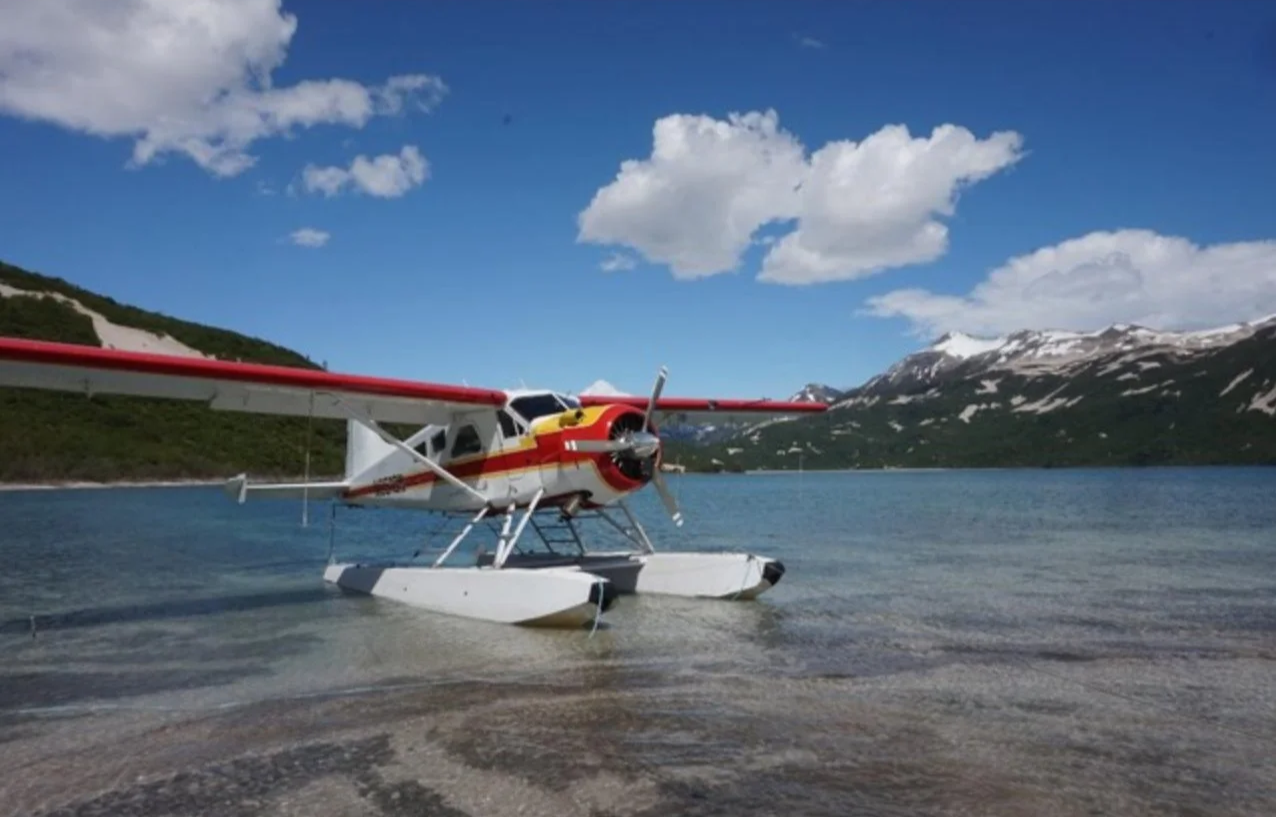 A seaplane with red and white colors parked on a lake with mountains in the background under a blue sky with scattered clouds.