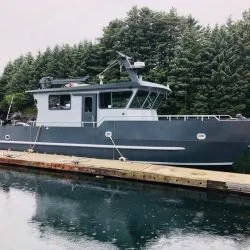 A gray military or patrol boat docked at a wooden pier on calm water with trees in the background.