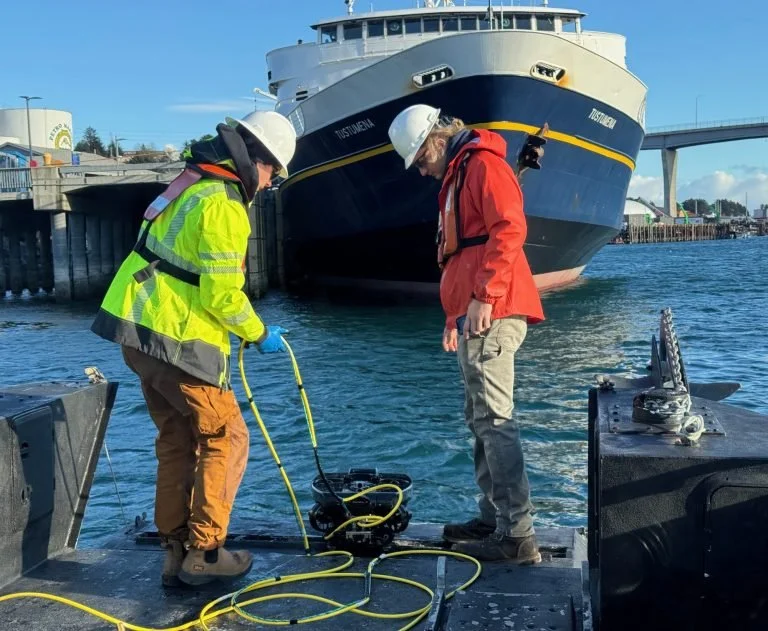 Two workers on the back of a boat preparing a small underwater robot with a large ship in the background, wearing safety helmets and jackets.