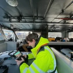 A person in a high-visibility yellow jacket using a handheld device inside a boat or ship control area
