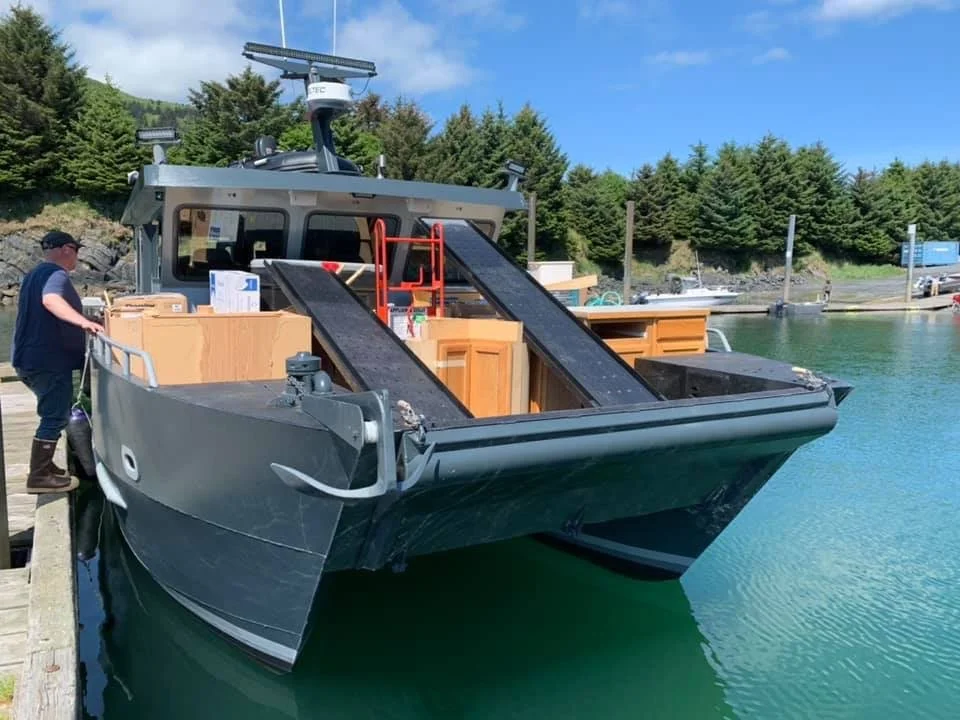 A person standing on a dock next to a large boat with a flat deck, wooden cabinets, and two inclined ramps.