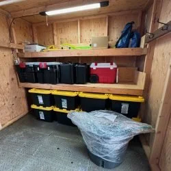 Storage shed interior with wooden shelves holding plastic bins and boxes, a workbench, and a garbage bag on the floor.