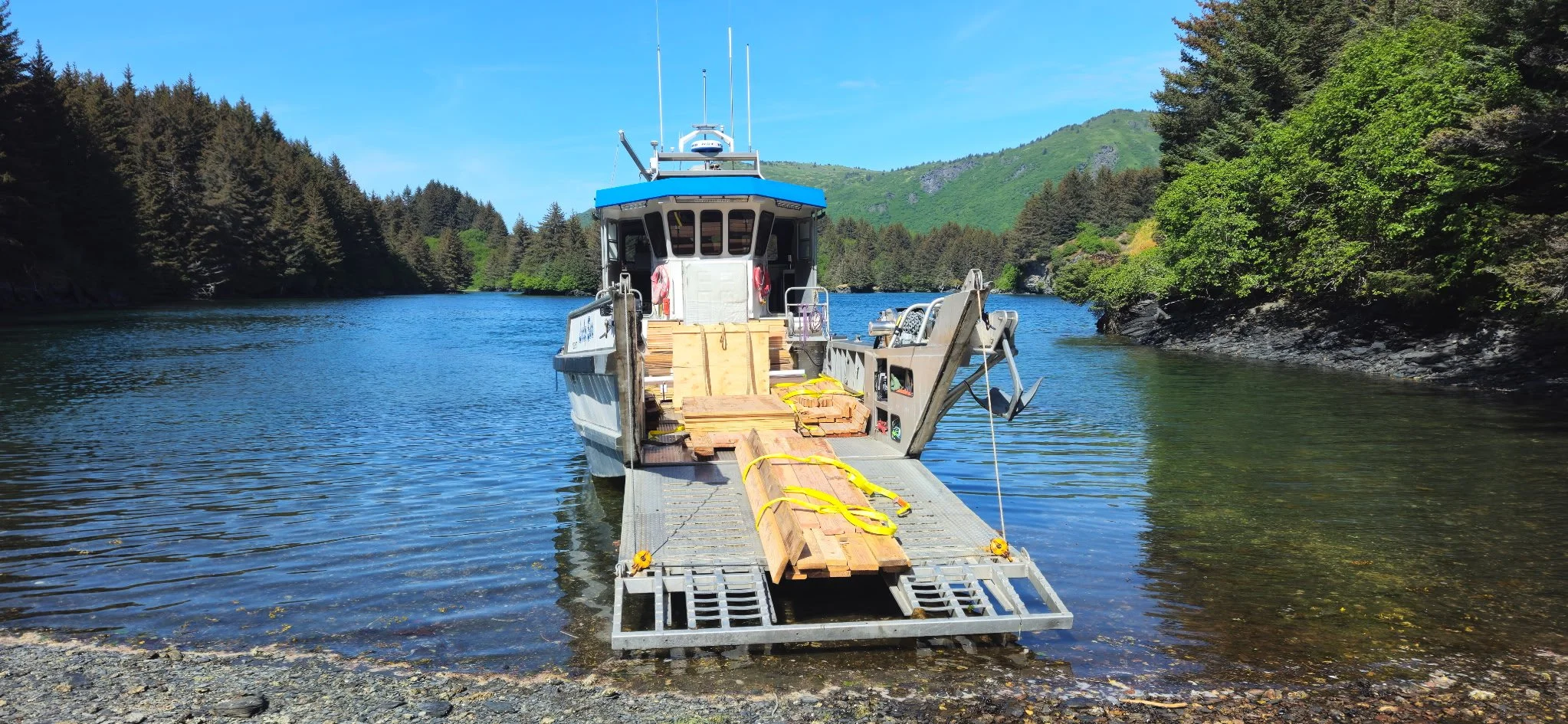 A boat docked at a lakeshore with wooden planks and straps on a ramp, surrounded by greenery and hills under a blue sky.