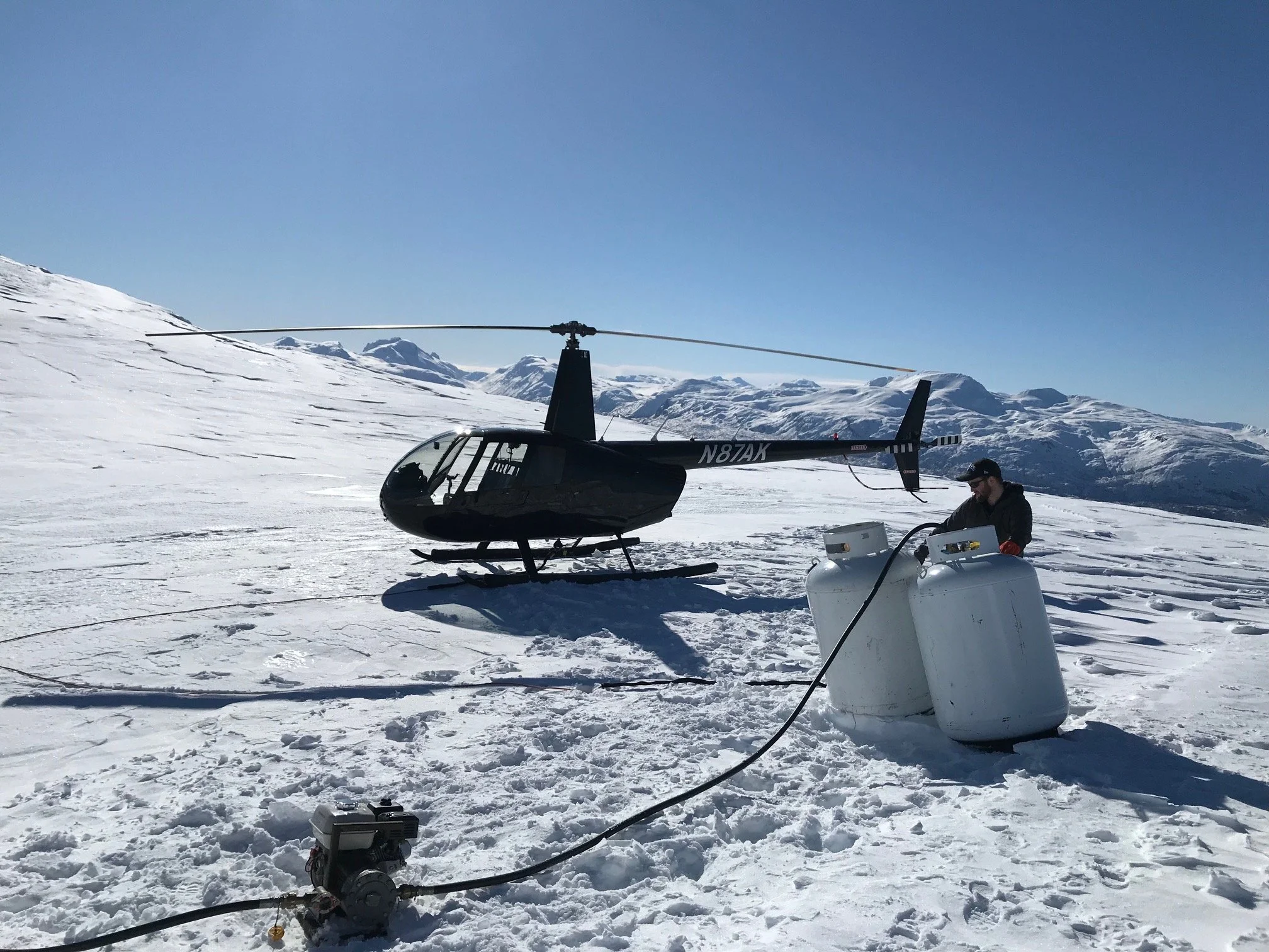 A person refueling a black helicopter on snowy mountain terrain with snow-covered mountains in the background.