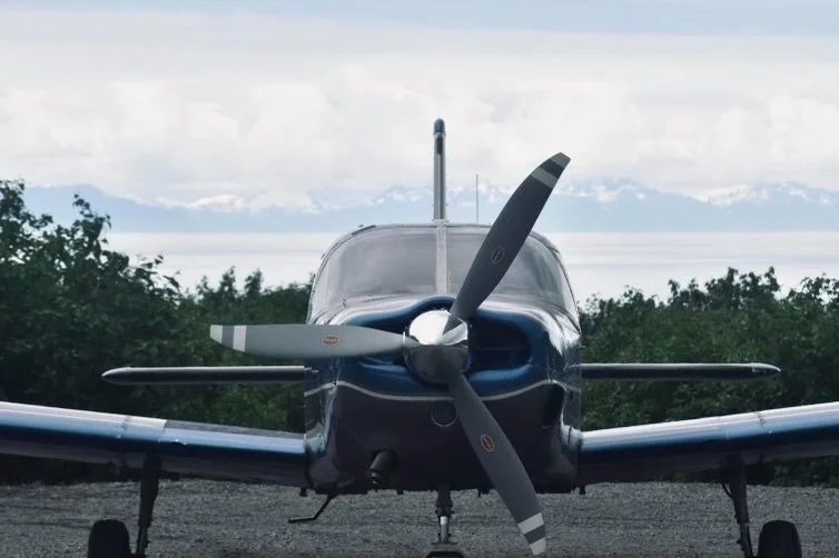 Front view of a small, single-engine aircraft with a two-blade propeller parked outdoors with trees and mountains in the background.