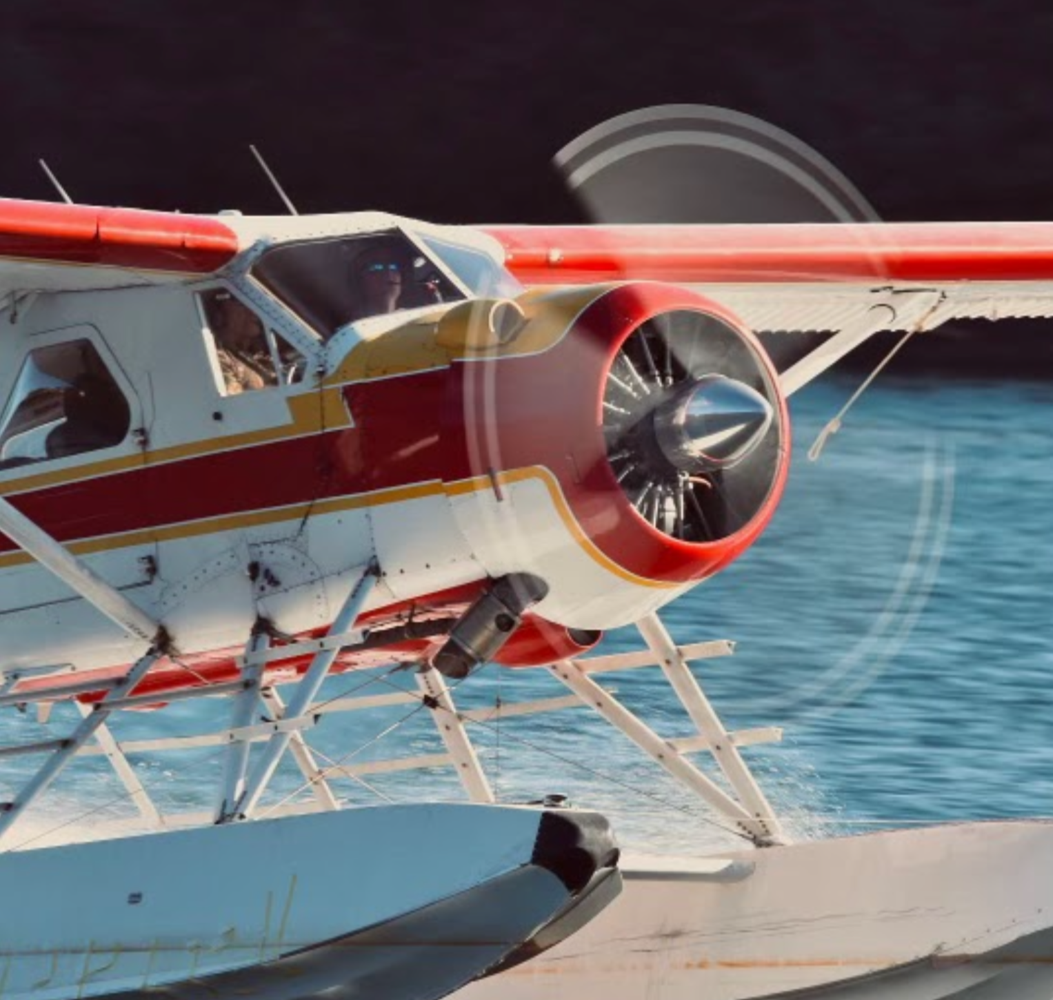 A seaplane flying over water, with the propeller visible in motion and the pilot visible through the cockpit window.