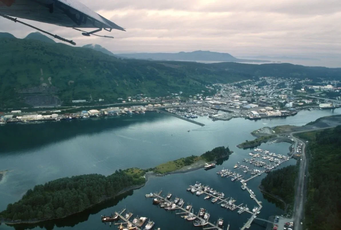 An aerial view of a harbor with boats docked, surrounded by water, green hills, and a city in the distance under a cloudy sky.