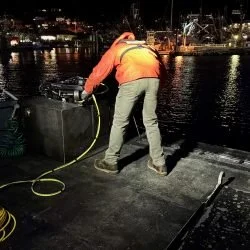A worker in an orange jacket and gray pants cleaning a dock at night with a hose, near a body of water illuminated by city lights.