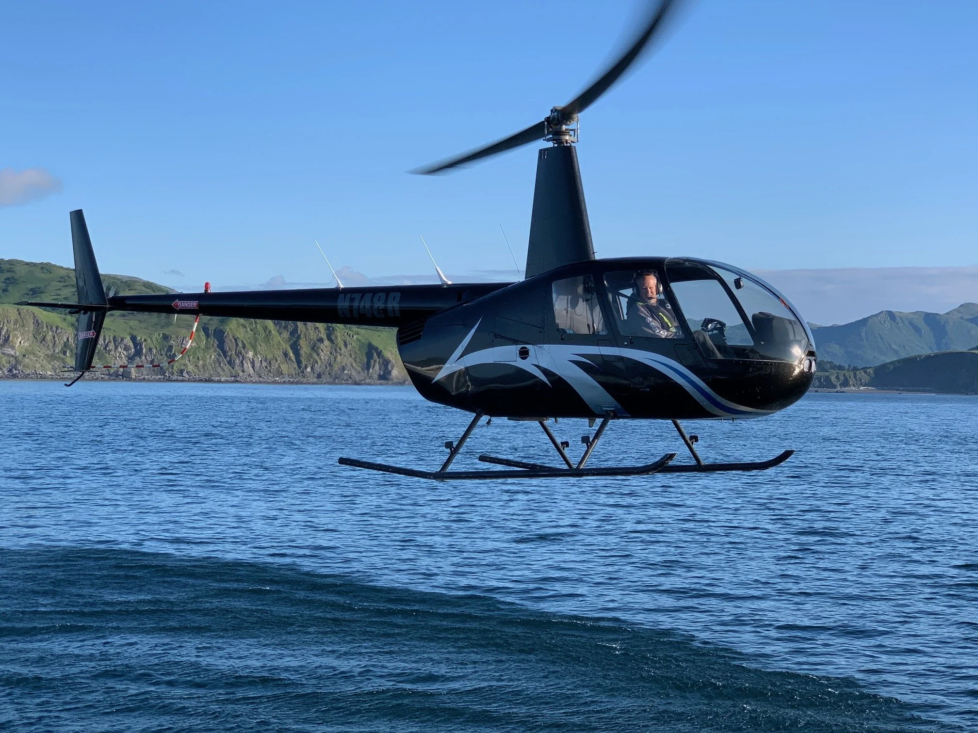 A black helicopter flying over a body of water with green hills and mountains in the background.