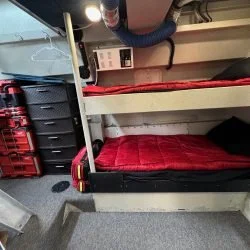Under a bunk bed with a red mattress, there is a black storage bin and a red plastic crate on the floor beside it, with a white wall and electrical outlet in the background.