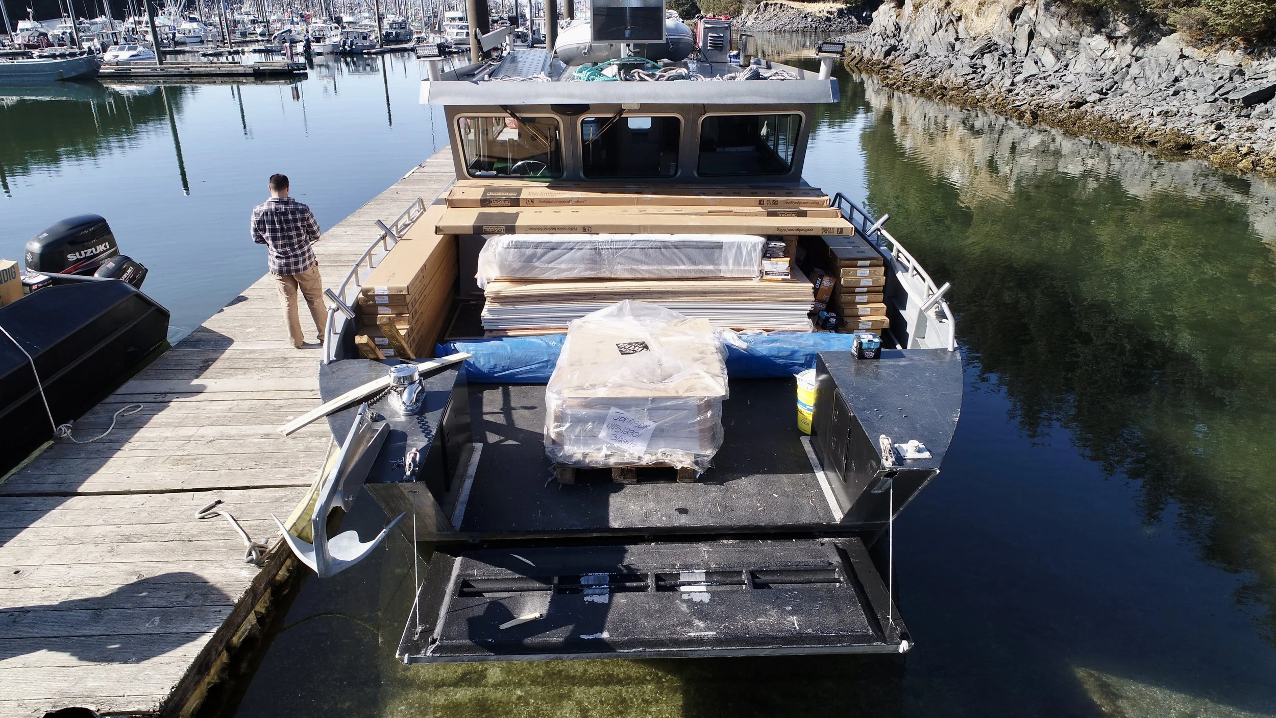 The image shows an empty boat docked at a marina with a man standing on the dock. The boat's deck is filled with stacks of wooden planks and packages covered in plastic. Other boats are docked nearby, and rocky terrain is visible along the shoreline.