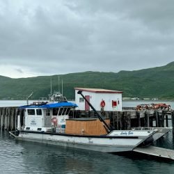 A docked boat with a small building on it, surrounded by water and green hills in the background under a cloudy sky.
