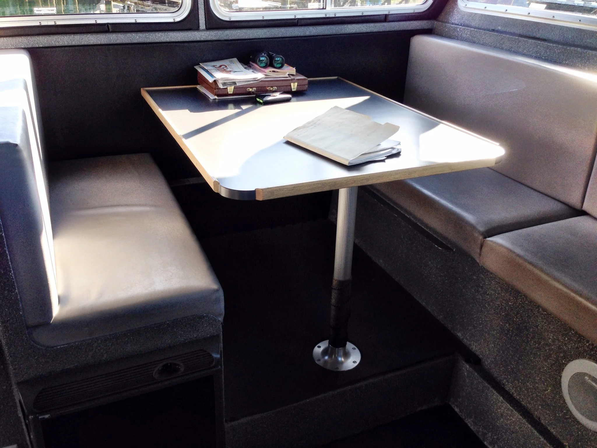 A booth in a train with a gray cushioned bench and a small rectangular table with papers, a notepad, and some small items on it. The windows show an outdoor scene with trees and sky.