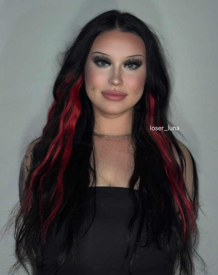 A woman with long black and red hair, wearing makeup, a black top, and a necklace, standing against a light background.