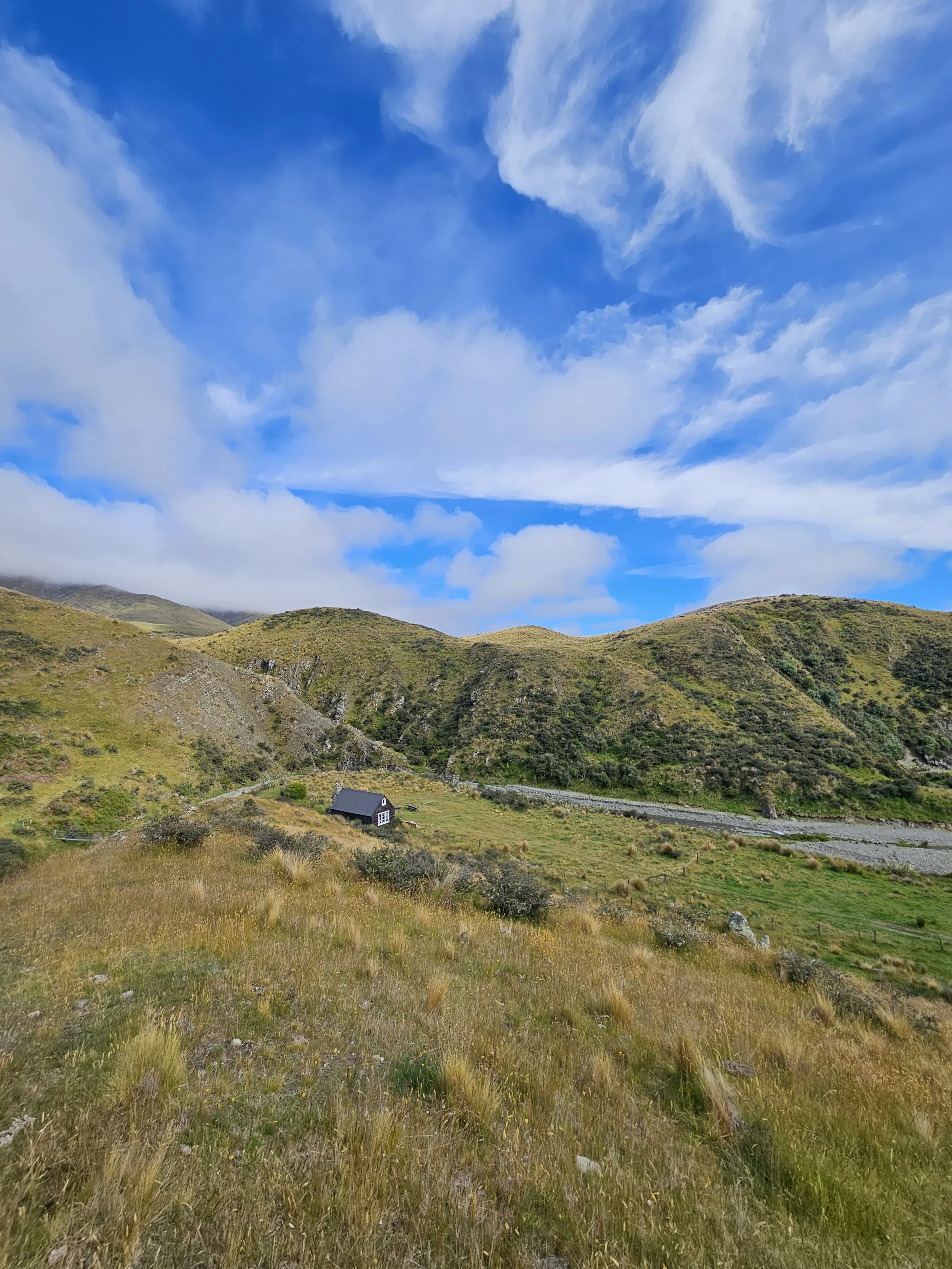 A small black house with a gray roof surrounded by green hills and a partly cloudy sky.
