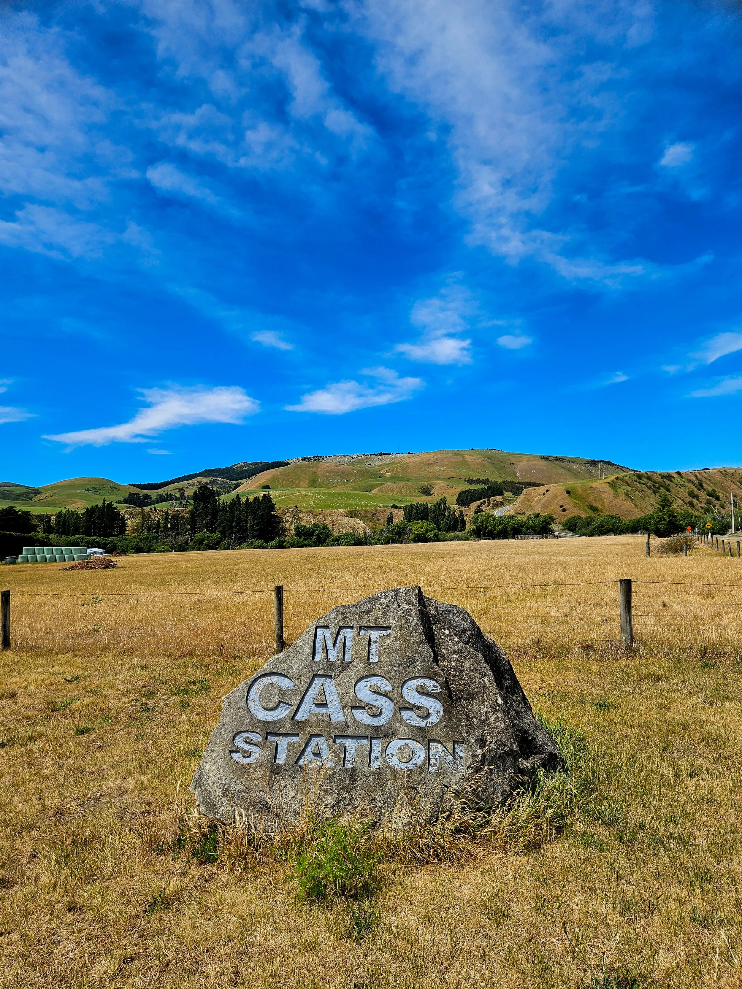 A large rock with the words 'Mt Cass Station' engraved on it, situated in a grassy field with a fence and rolling hills in the background under a blue sky with scattered clouds.