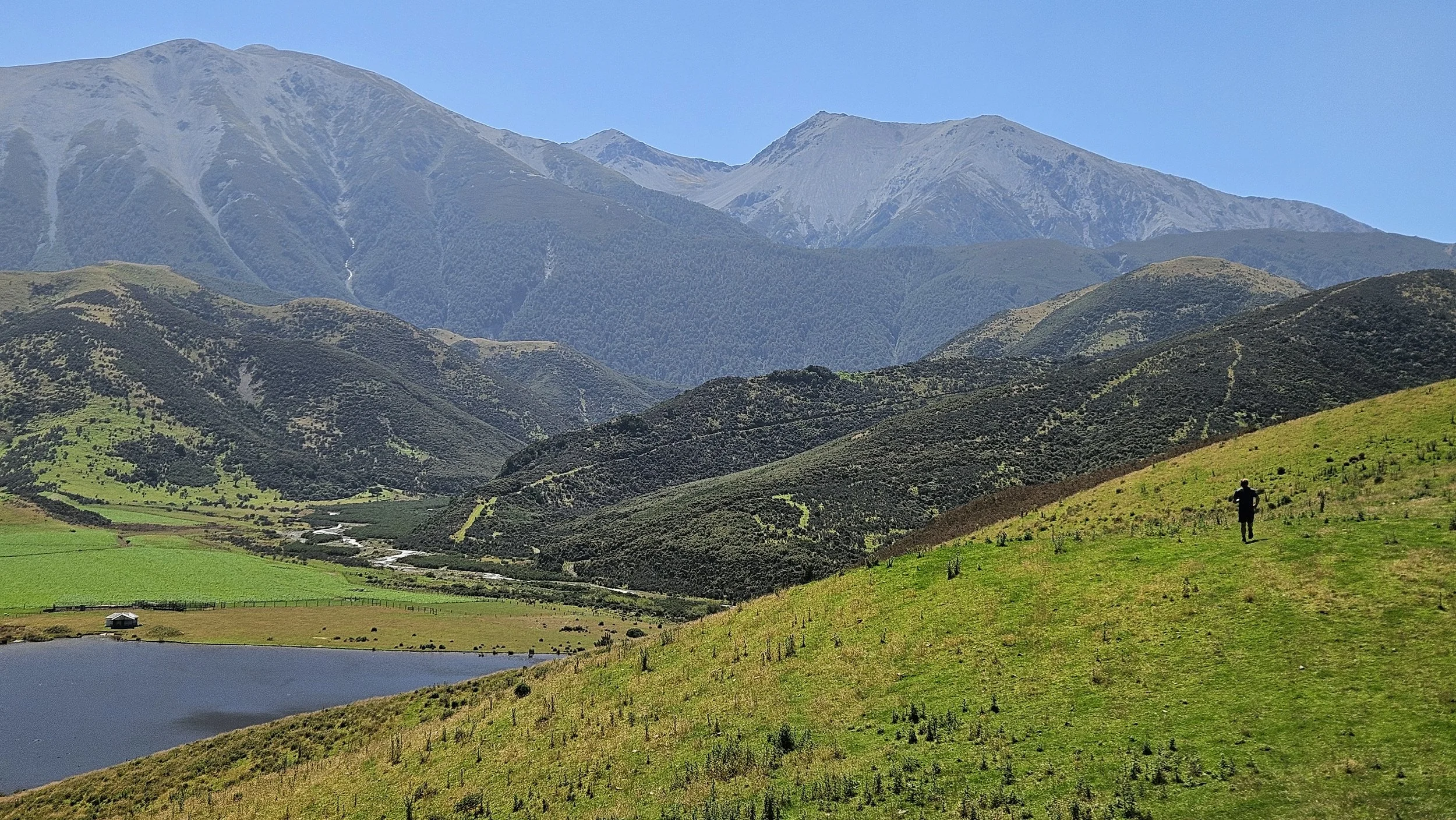 A person walking on a grassy hill with a small pond nearby, surrounded by rolling green hills and distant mountains under a clear blue sky.