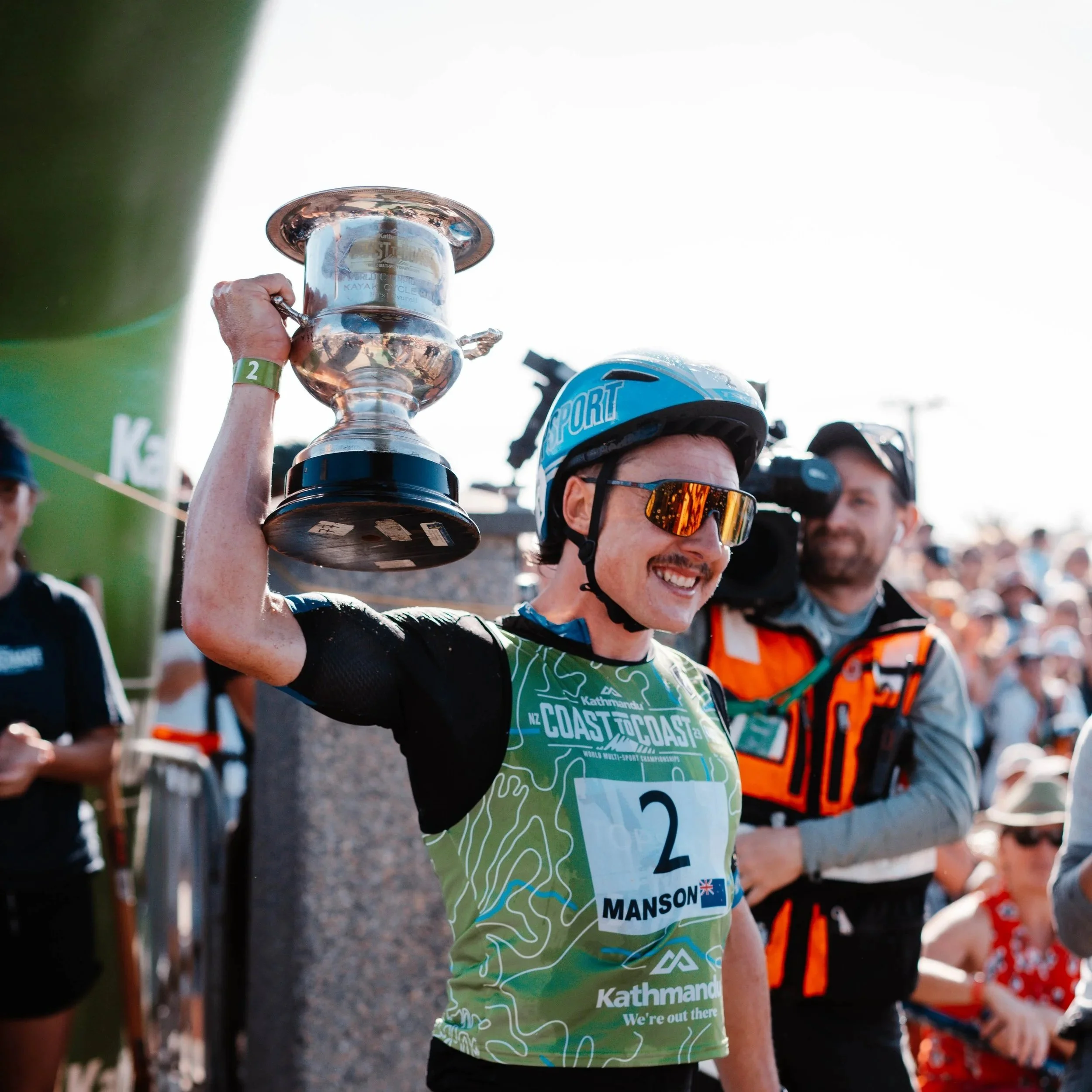 A cyclist wearing a helmet and sunglasses, holding a large silver trophy above his head, smiling at a cycling event with people and a green arch in the background.
