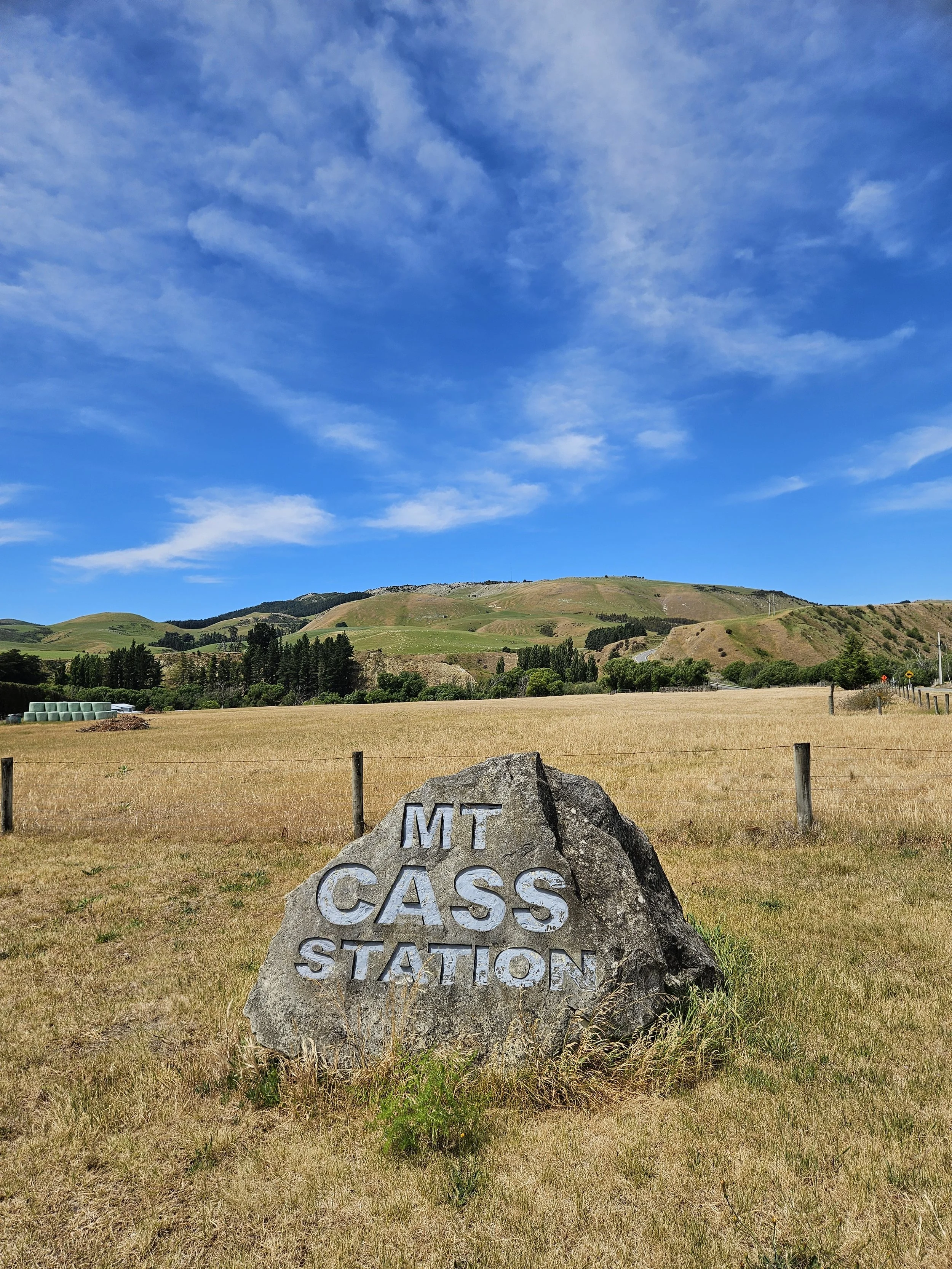 A large sign carved into a rock with the words 'MT CASS STATION' on a grassy field with mountains and a blue sky in the background.