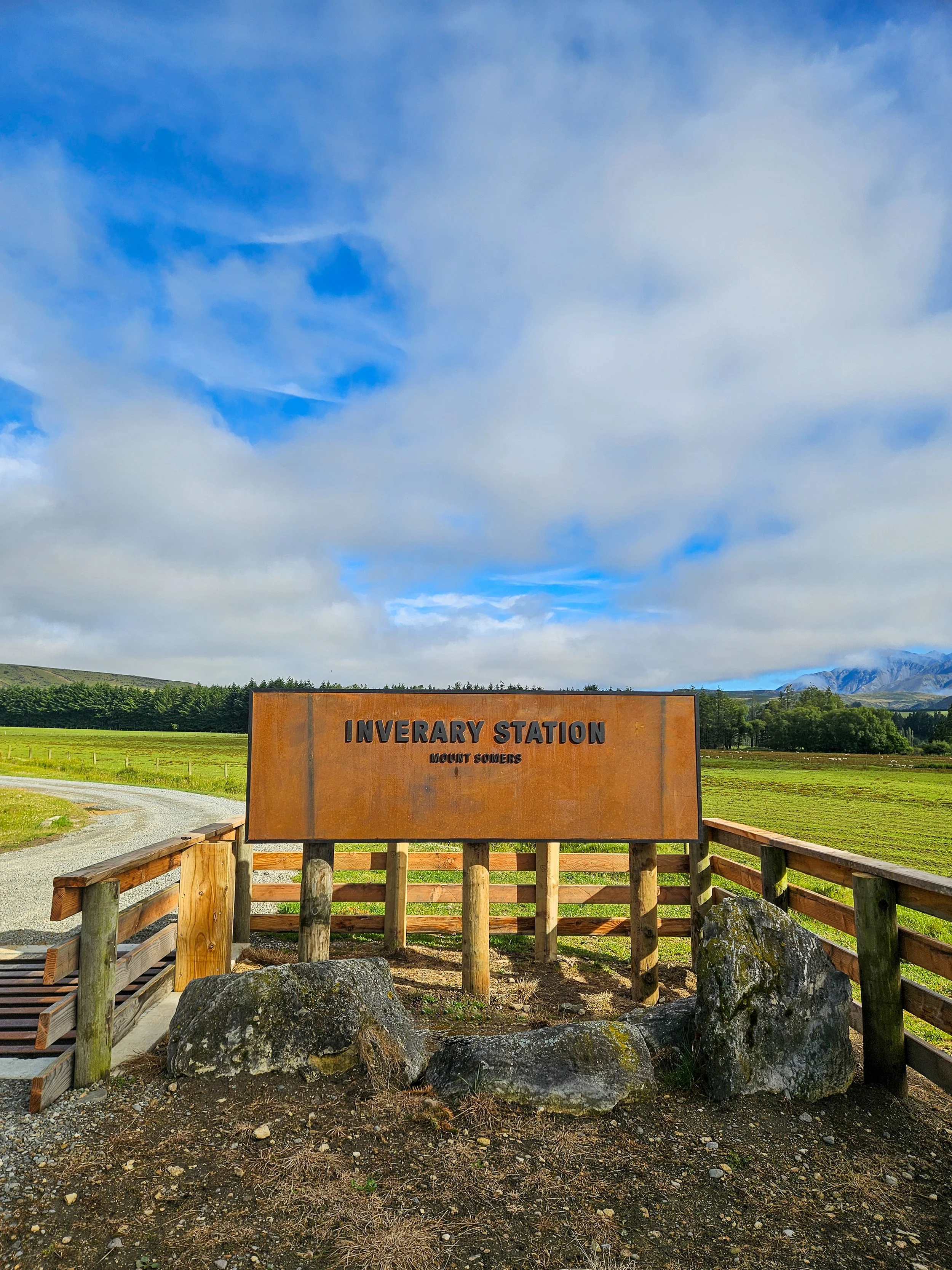 A sign at Inverary Station, Mount Somers, in a rural, grassy landscape with a gravel path, wooden rails, rocks, trees, and mountains in the background under a partly cloudy sky.