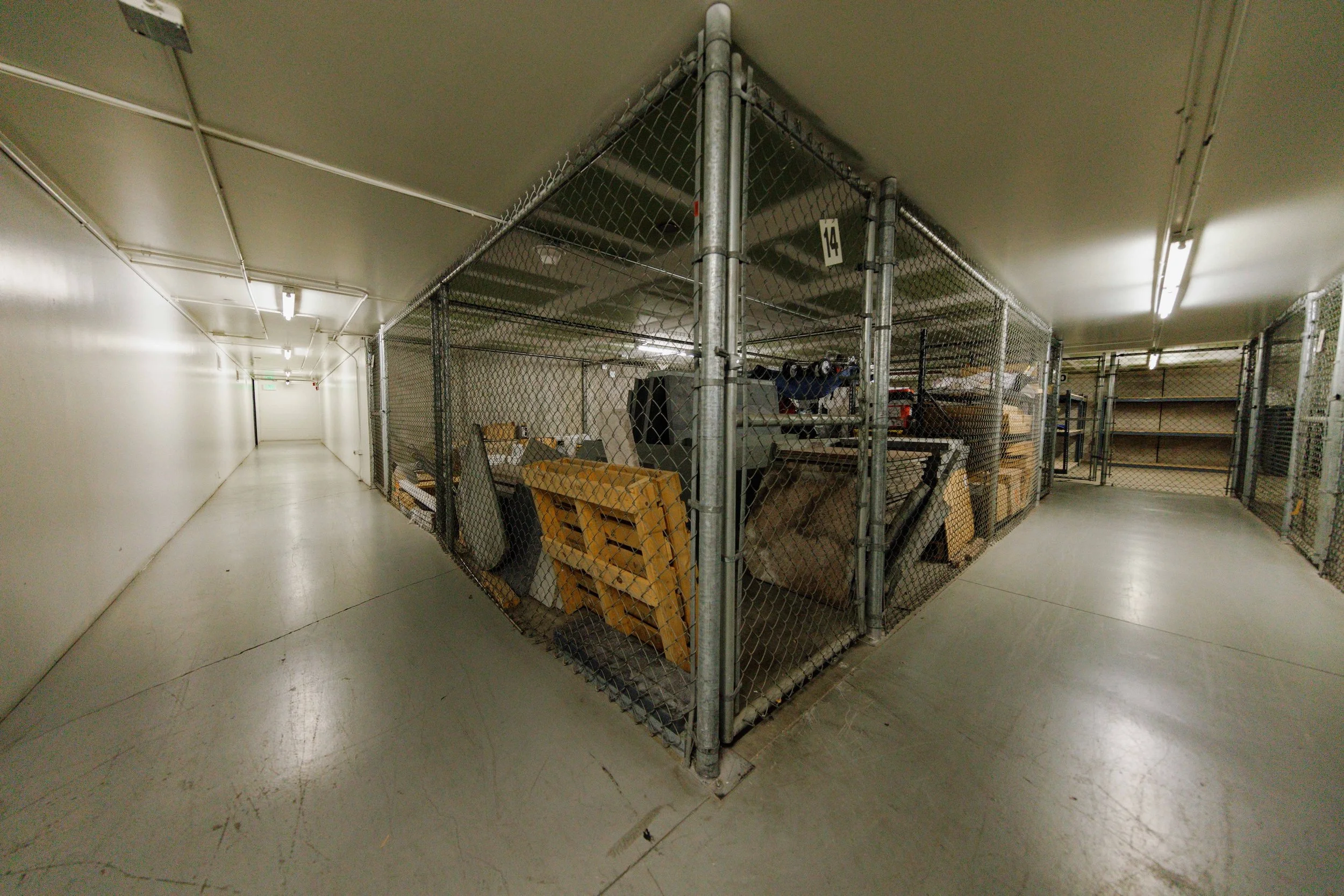 Underground storage area with chained cages containing pallets, boxes, and equipment, in a well-lit concrete corridor.