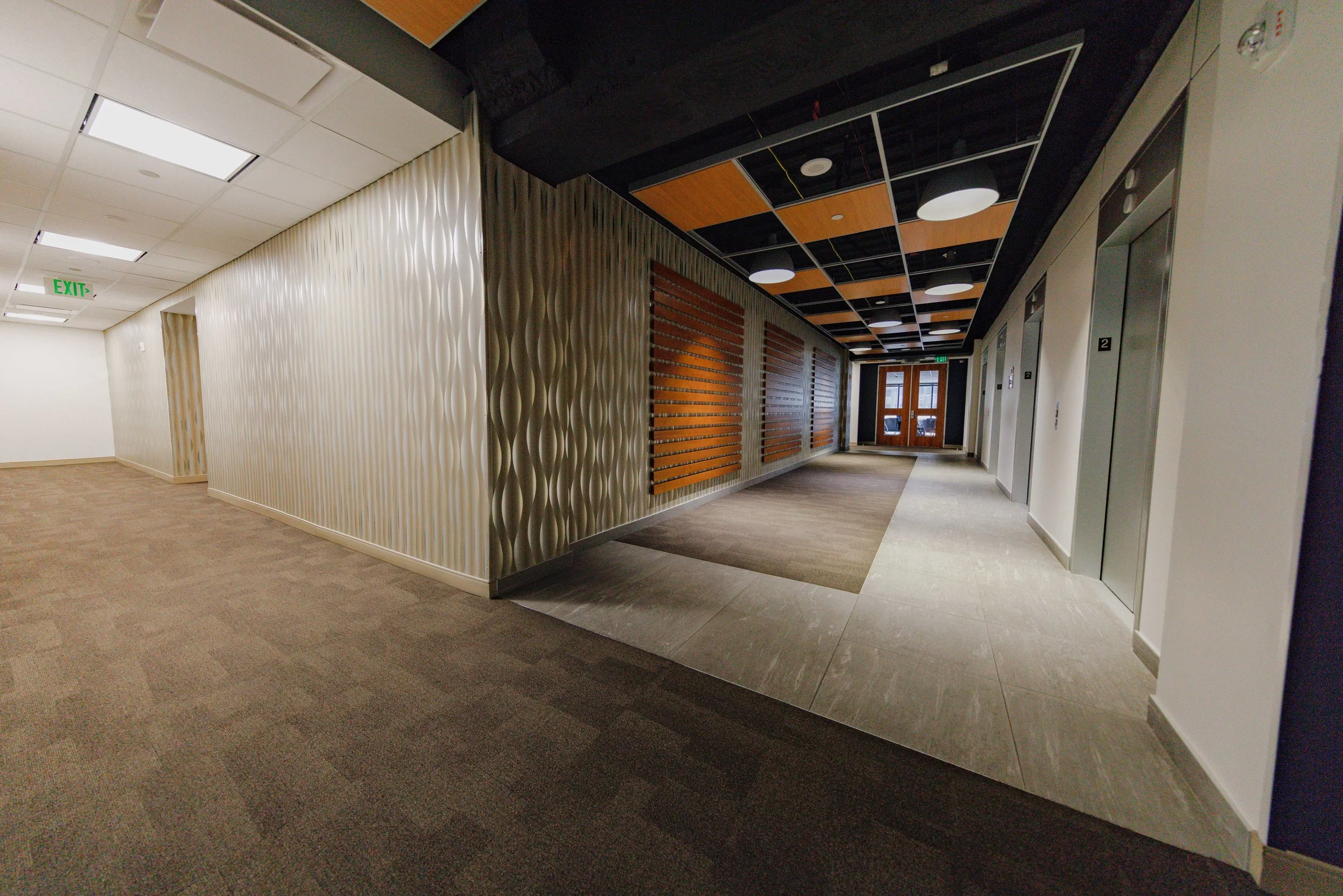 Modern hallway with elevator doors, decorative wall panels, and wooden accents, leading to glass double doors at the end.