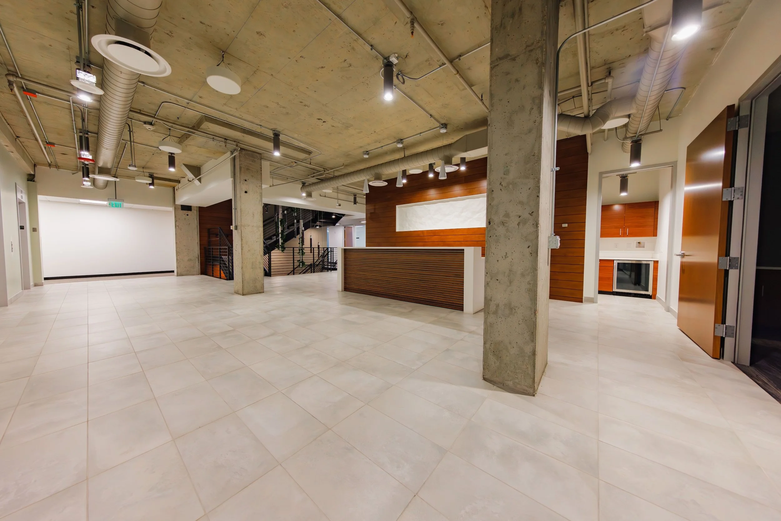 Empty modern lobby or reception area with tiled floor, exposed concrete ceiling with visible ductwork, wooden accents, and staircase in background.