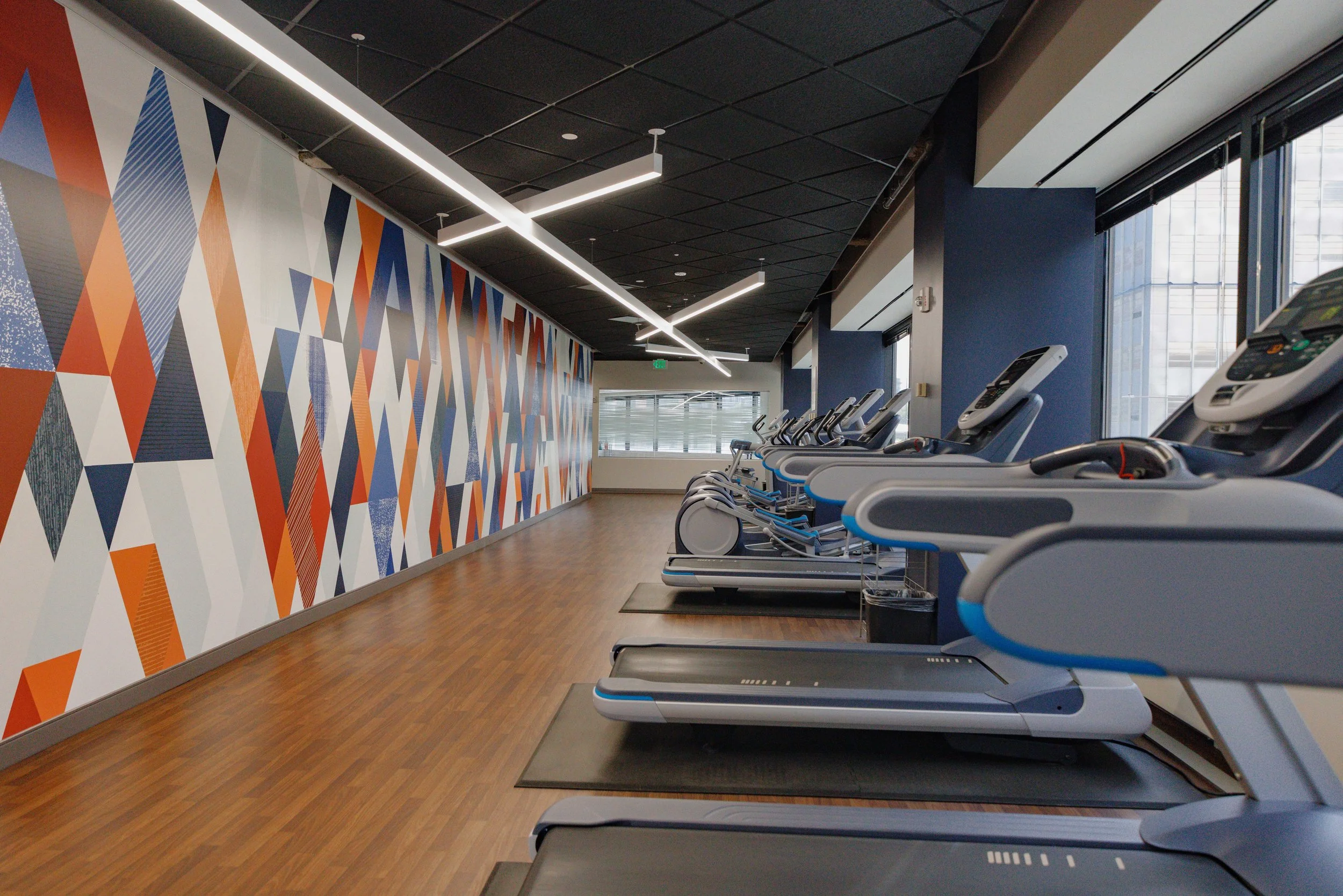 Empty gym with multiple treadmills lined up near a large window, colorful geometric wall art, and modern lighting fixtures.