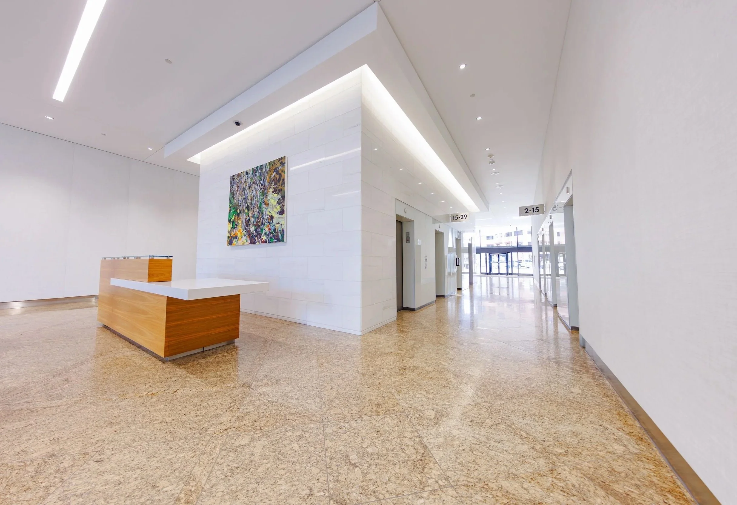 Lobby of a modern building with white walls, a wooden reception desk, and artwork on the wall. There are elevators and signs indicating floor ranges, and glass doors at the far end letting in natural light.