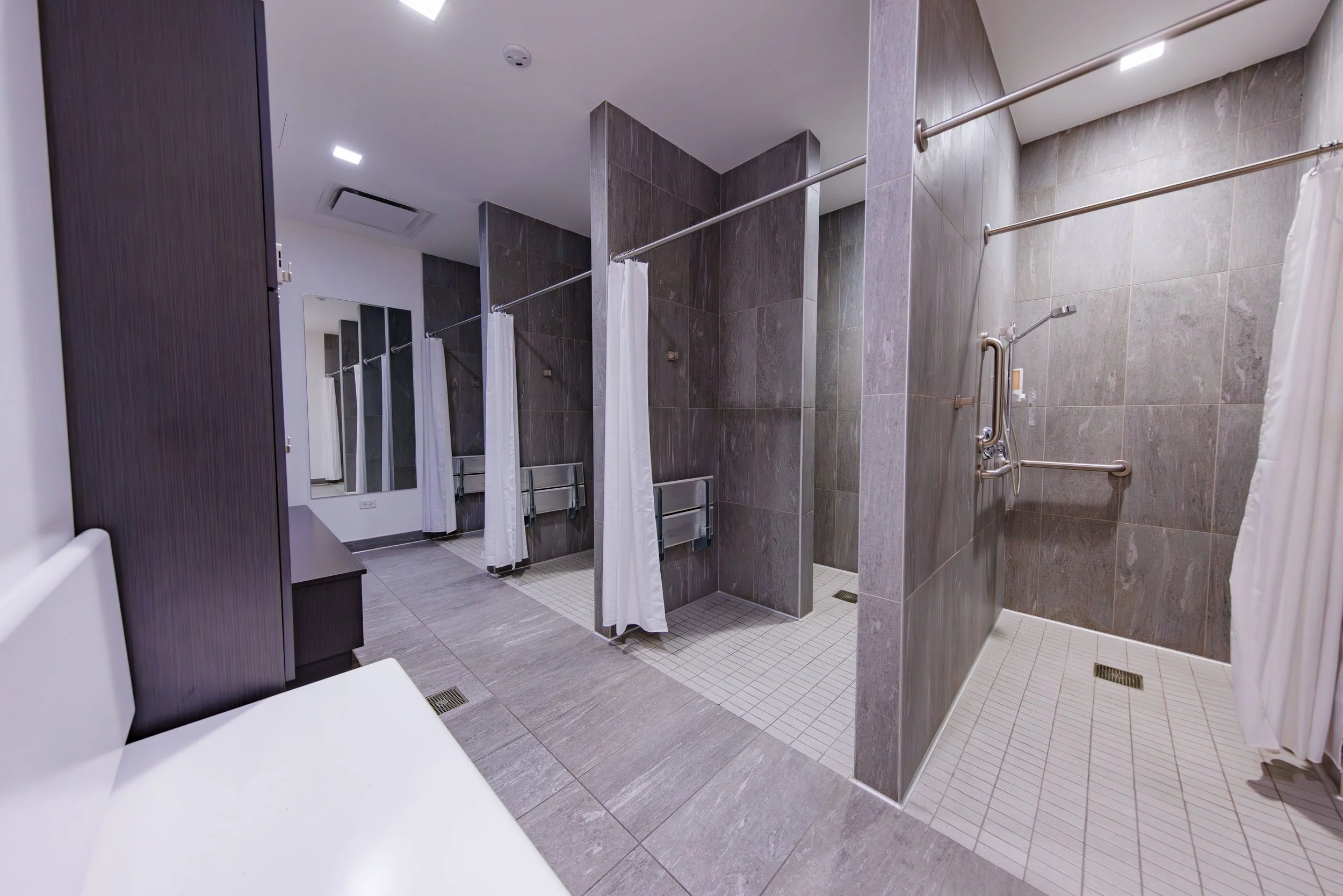 Men's accessible shower rooms with individual stalls, bars, and showerheads in a modern, clean, and well-lit bathroom.