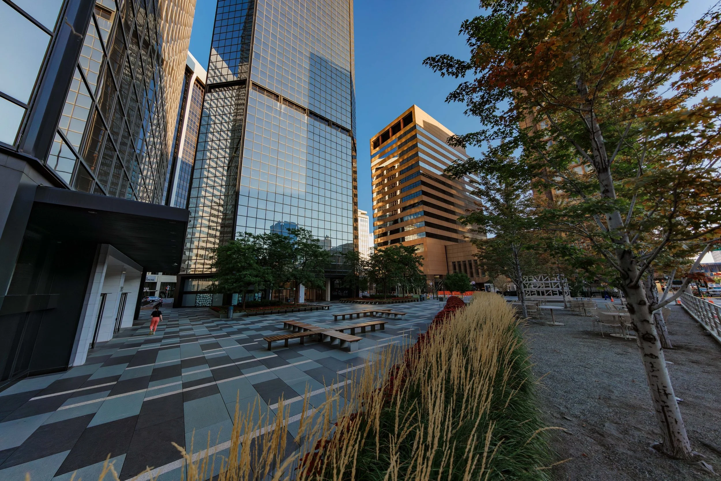 Urban park area surrounded by tall modern glass buildings with benches, trees, and grass, during daytime.