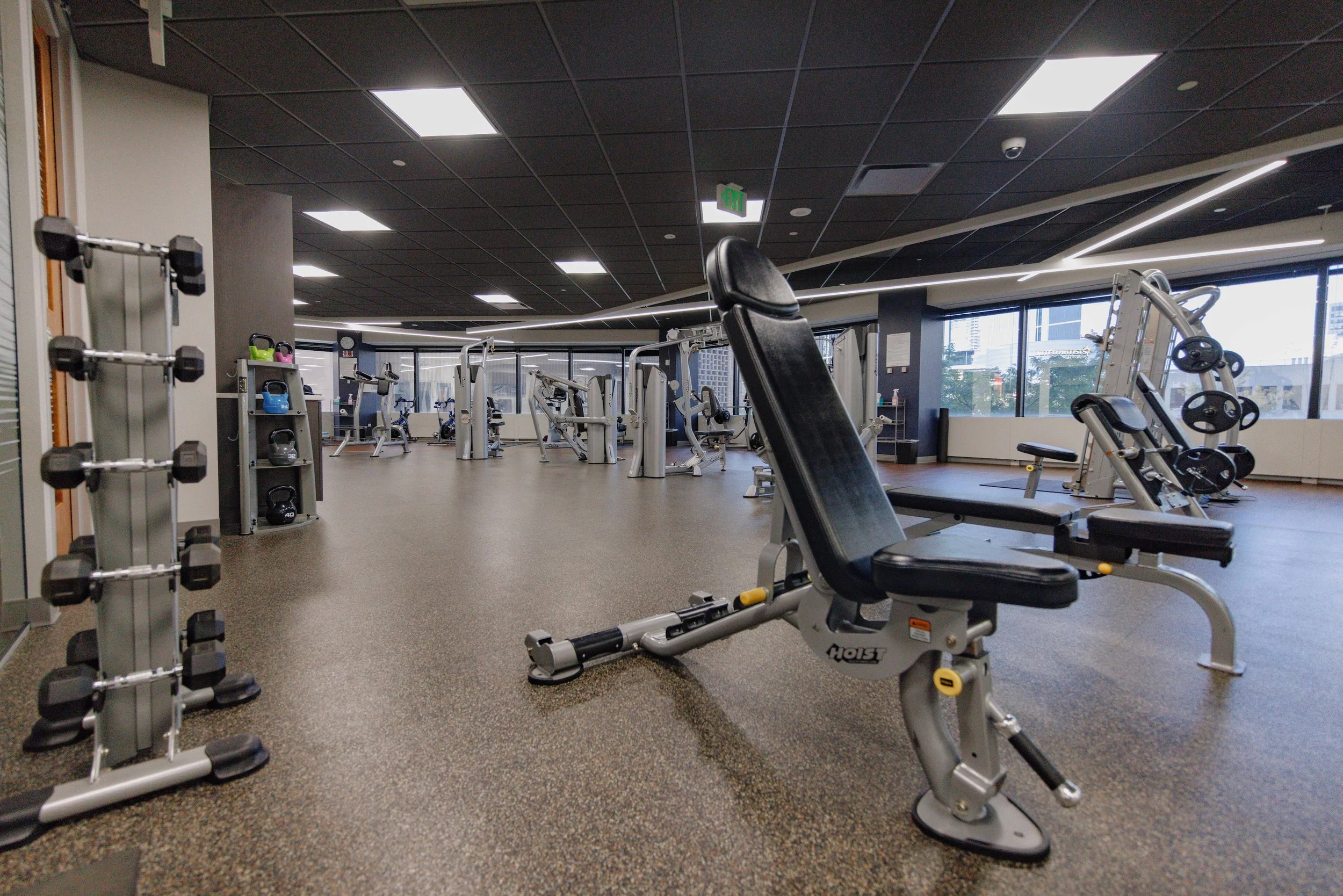 Empty gym with workout equipment including a weight rack, benches, and cardio machines, with large windows showing city buildings outside.