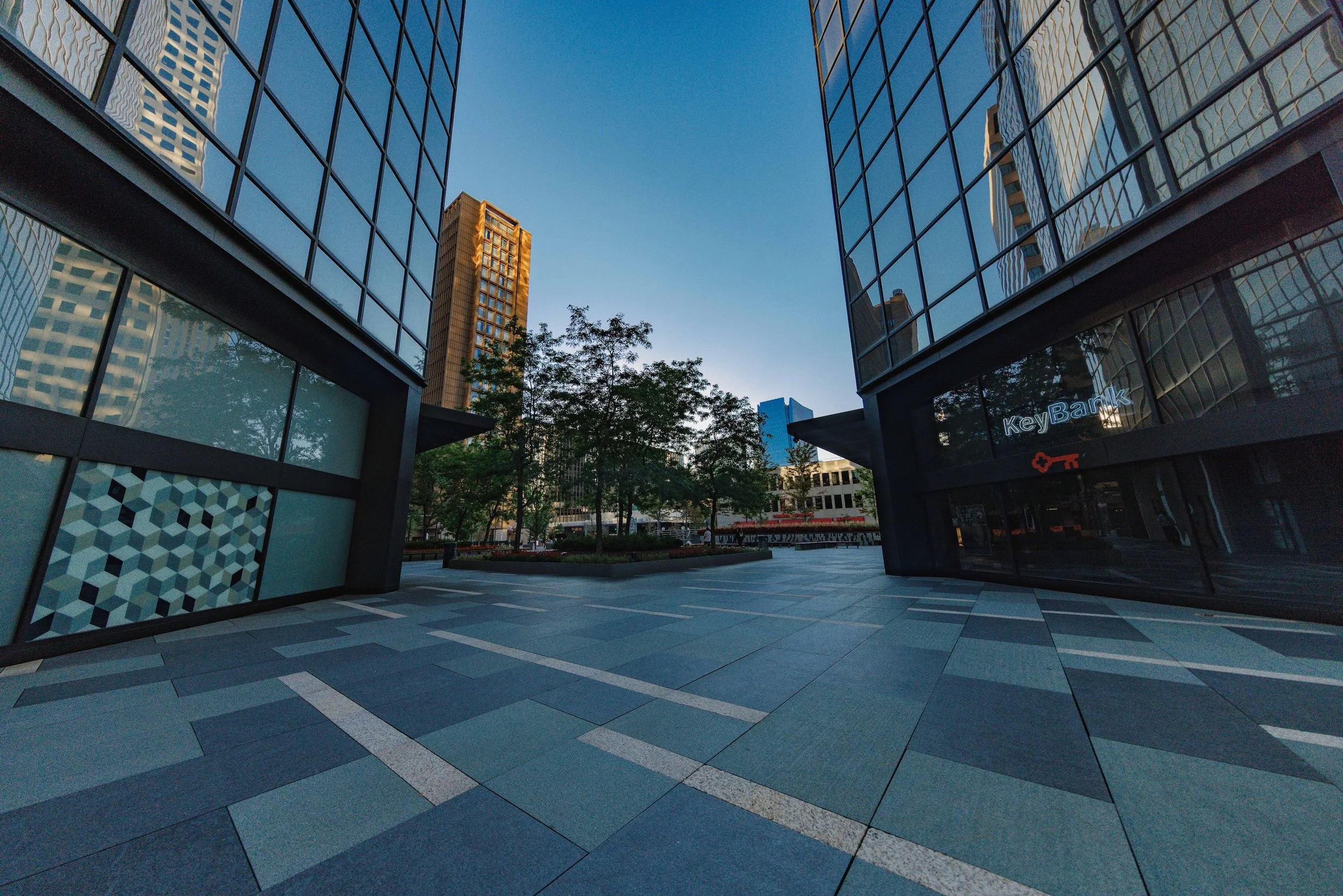 Empty outdoor plaza between modern glass office buildings with trees and a clear blue sky.