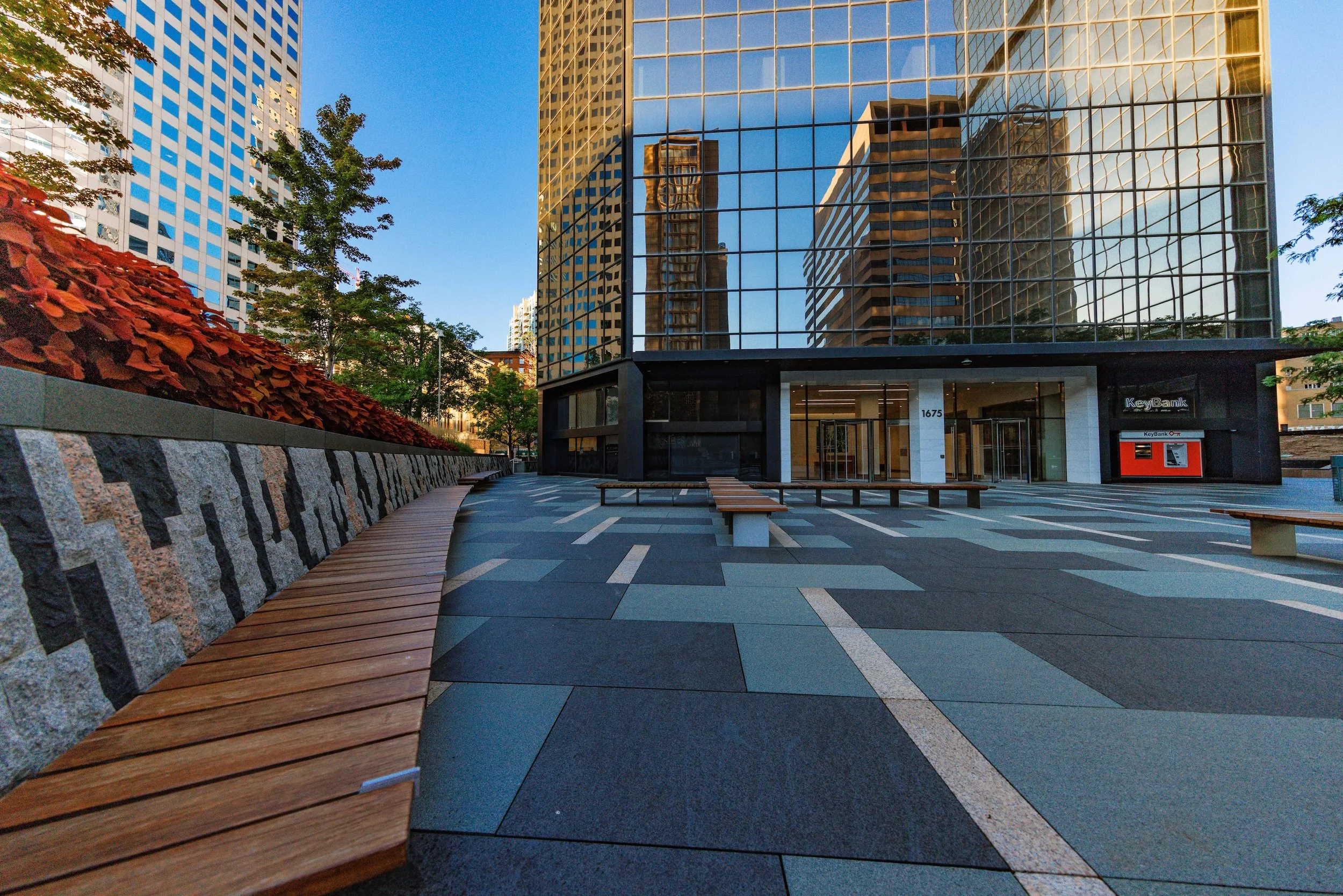Empty urban plaza with patterned paving and benches, surrounded by tall modern glass buildings reflecting the blue sky, with trees and a stone wall with wooden seating on the left.