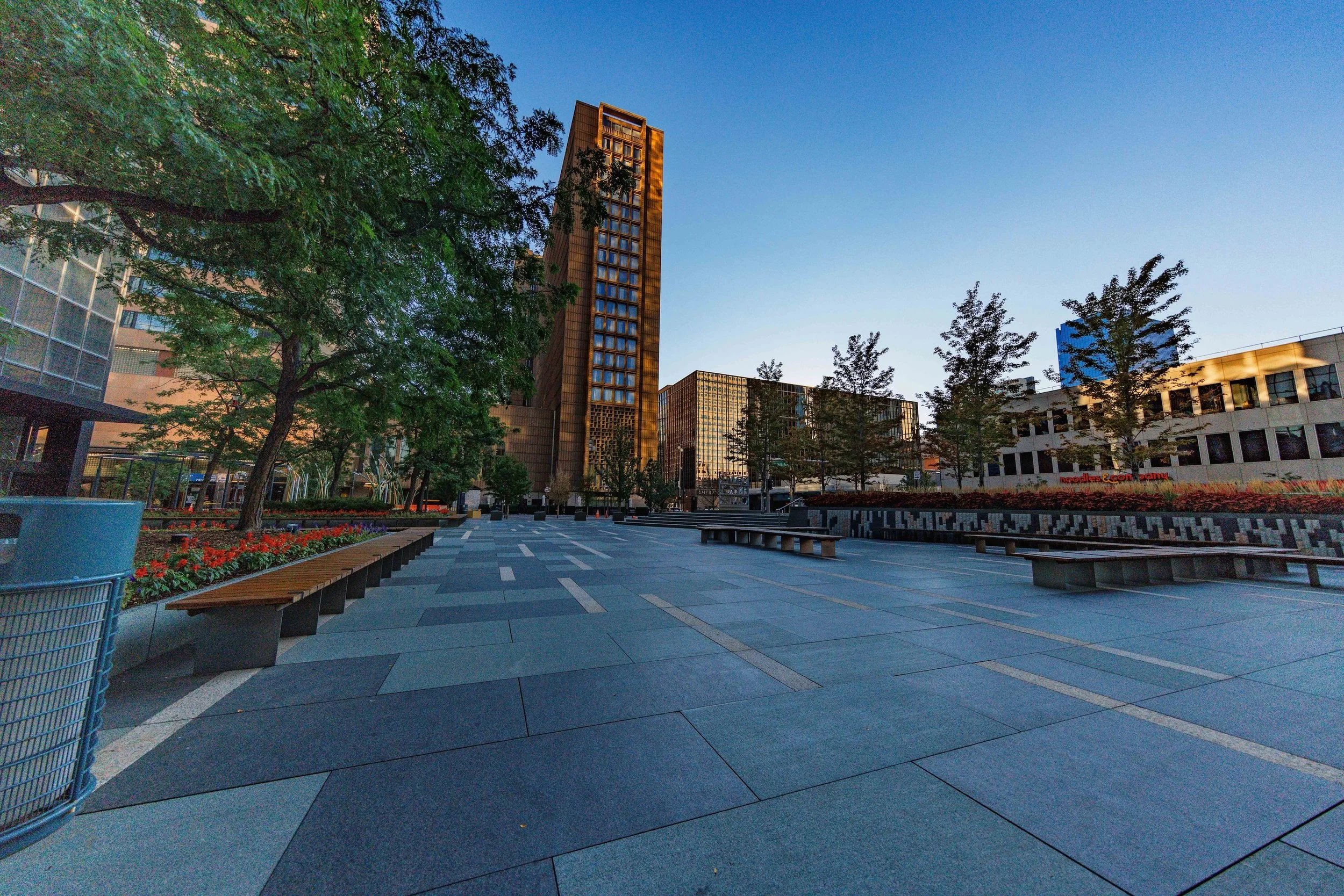 An empty urban plaza with modern buildings, trees, benches, and flower beds under a clear blue sky.
