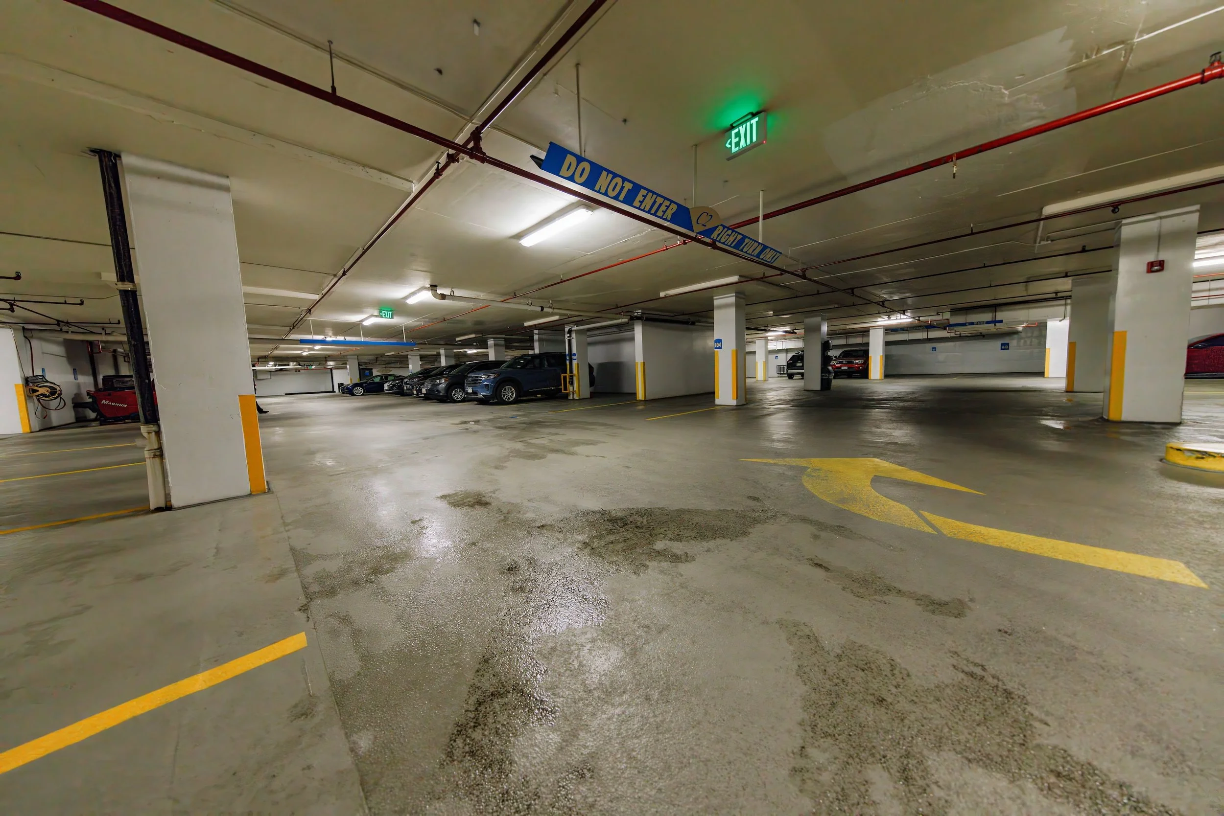 Underground parking garage with several parked cars, yellow lines, and directional arrows on wet concrete floor. Exit sign and a blue sign that says 'Do Not Enter' are visible on ceiling.