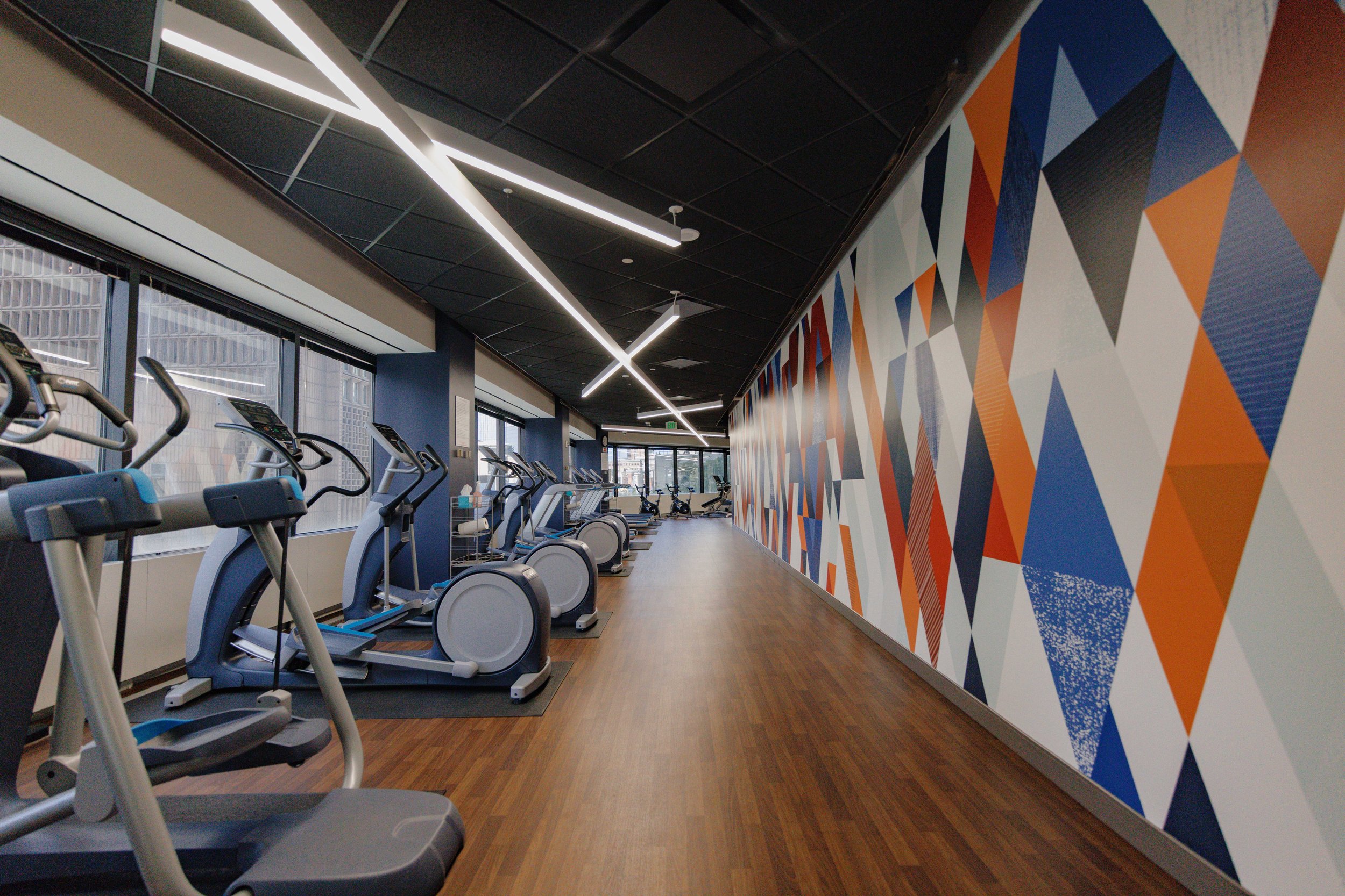 Empty gym with exercise machines lined up near large windows and a colorful geometric mural on the wall.