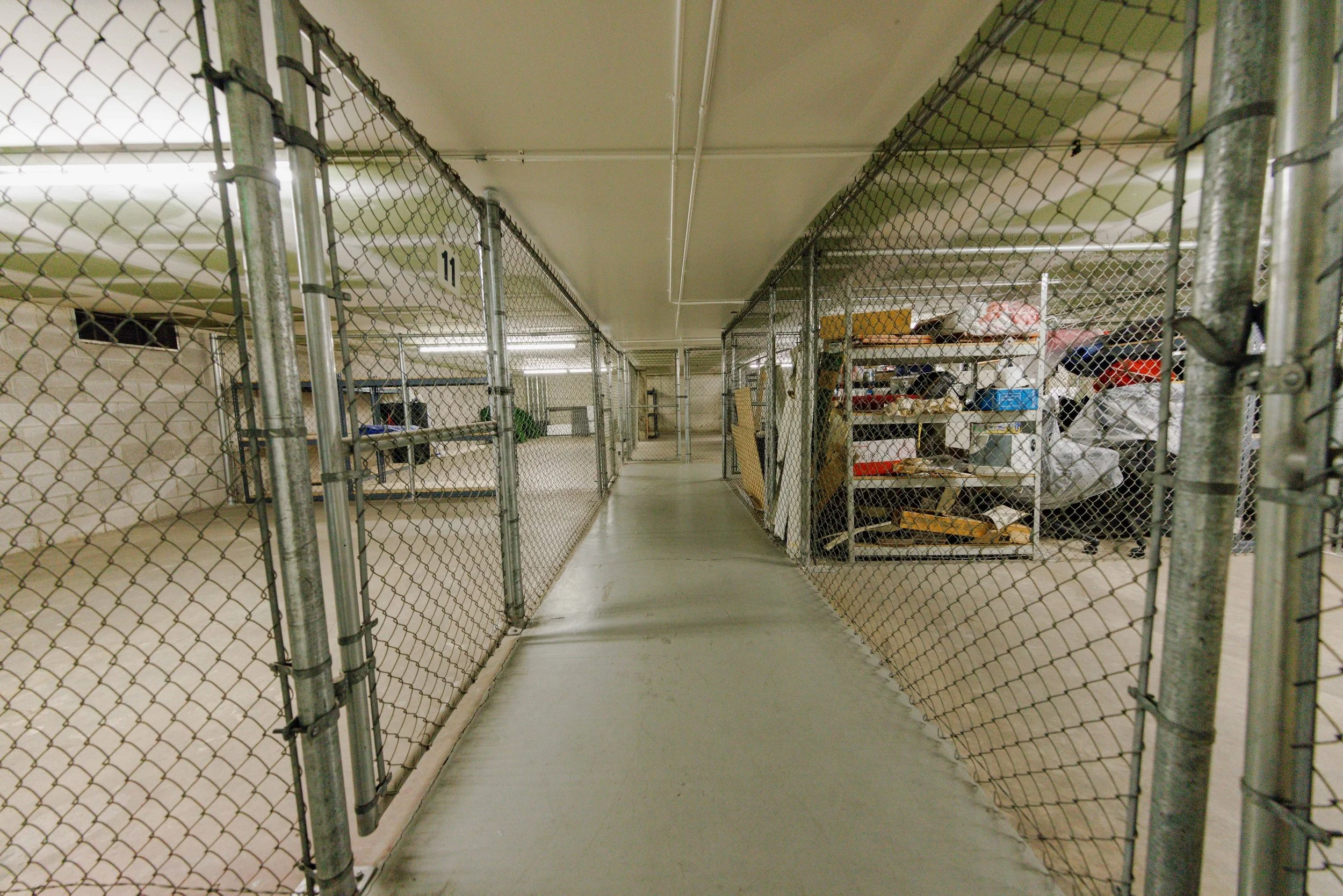 Empty storage unit with chain-link fence door, shelves filled with miscellaneous items, in a warehouse.